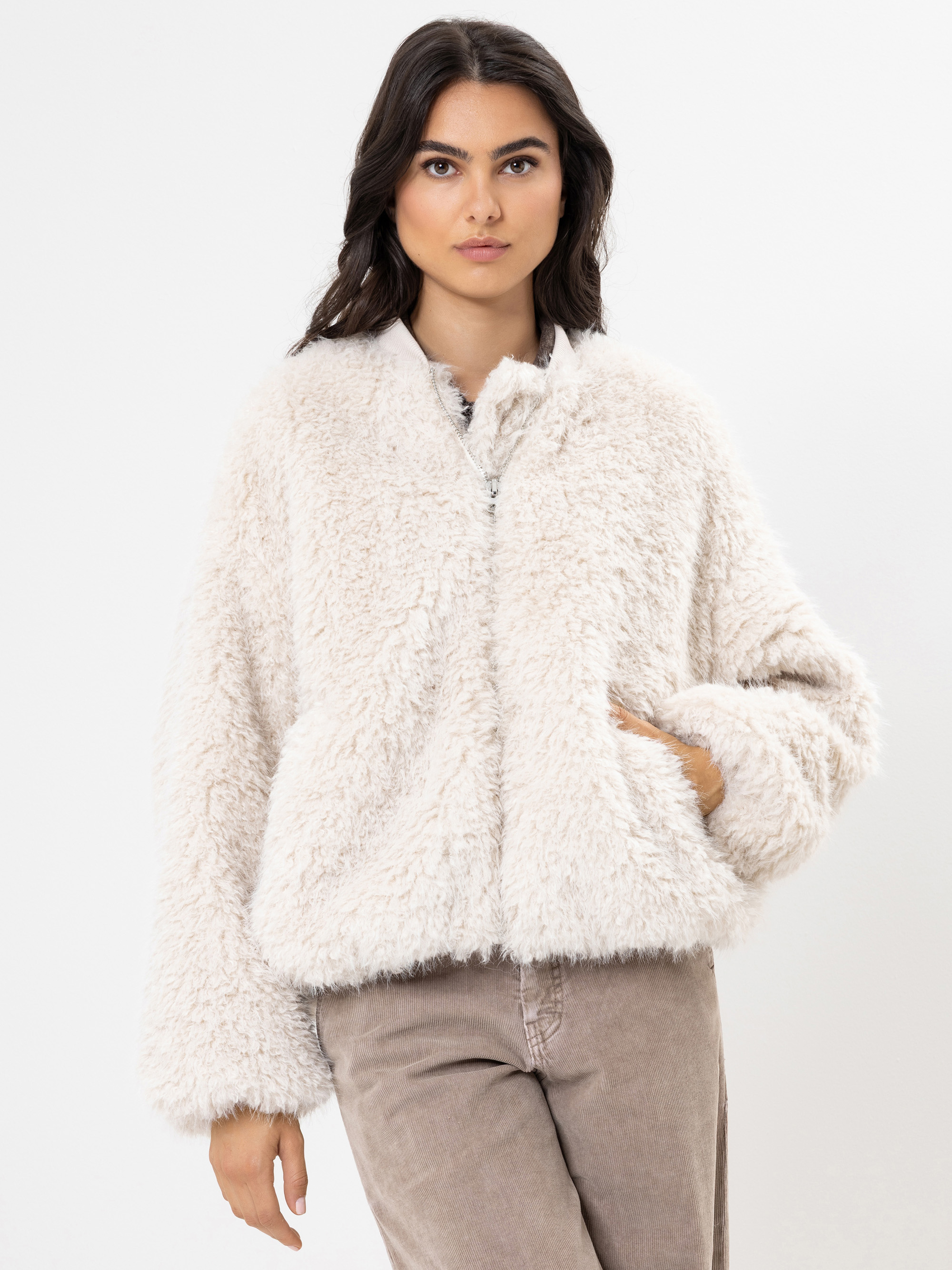 Eine Frau, die eine flauschige, cremefarbene Kunstpelzjacke mit der linken Hand in der Tasche trägt, kombiniert mit einer hellbraunen Hose, steht vor einem schlichten weißen Hintergrund.