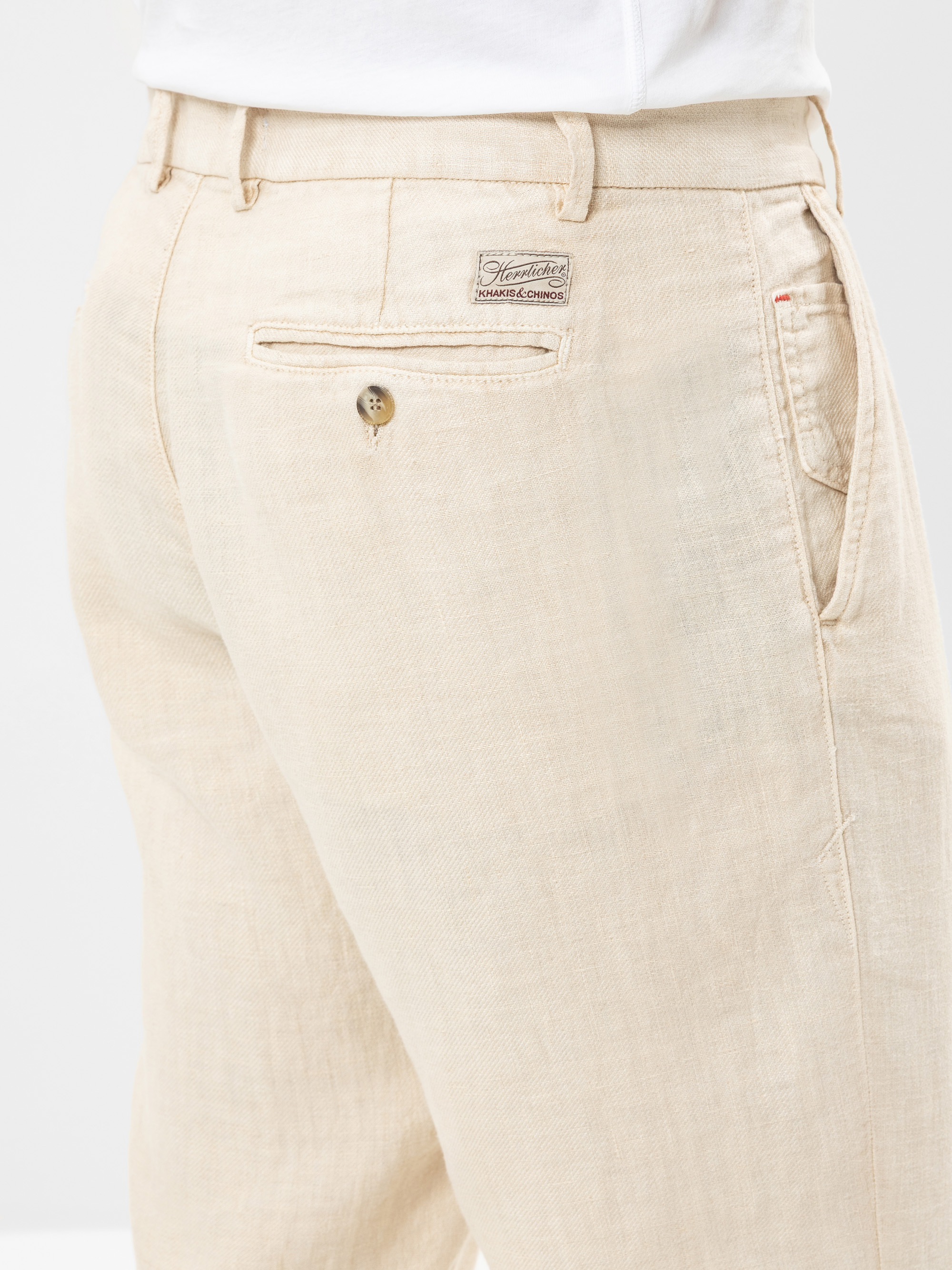 Nahaufnahme einer Person, die eine hellbeige Leinenhose trägt, wobei der Schwerpunkt auf dem Bereich der rechten Gesäßtasche mit einem Knopf und einem aufgenähten Markenetikett über der Tasche liegt. Die Hose hat eine lockere Passform und eine sichtb
