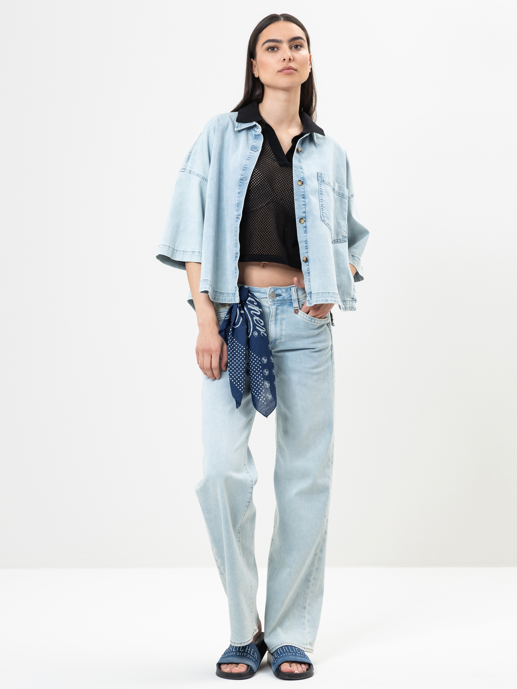 Eine Frau steht vor einem schlichten Hintergrund und trägt eine hellblaue Jeansjacke, dazu passende weite Jeans, ein schwarzes Crop-Top, ein blaues Kopftuch, das sie um die Taille gebunden hat, und blaue Sandalen. Sie hat langes dunkles Haar und einen ne