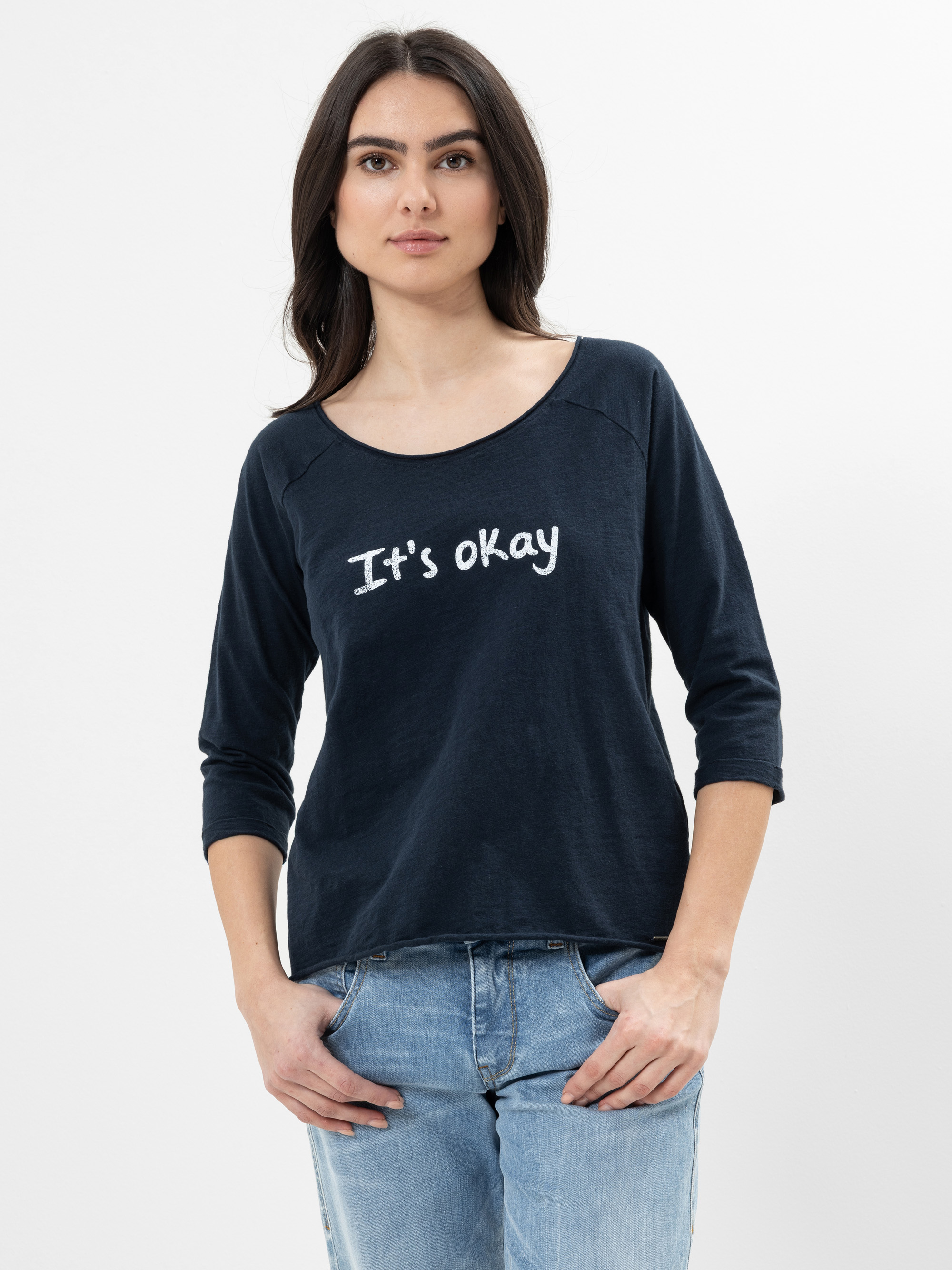 Eine Frau mit langen dunklen Haaren, die ein marineblaues Hemd mit der Aufschrift Its okay in weißer Schrift trägt, steht mit den Händen in den Taschen vor einem schlichten weißen Hintergrund.