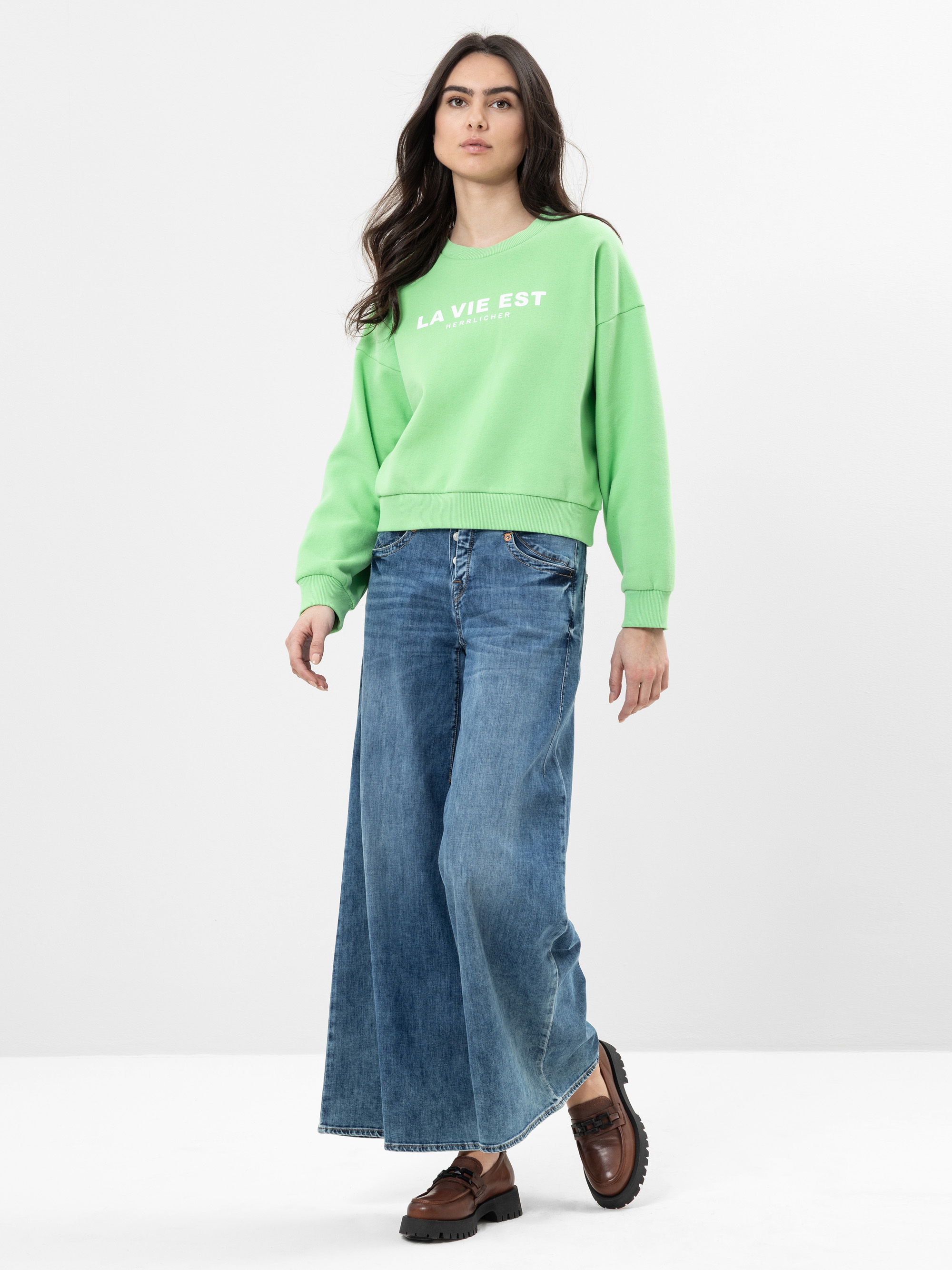 Eine Frau steht vor einem schlichten Hintergrund und trägt ein hellgrünes Sweatshirt mit weißer Aufschrift und eine weit geschnittene blaue Jeans. Sie hat langes dunkles Haar und trägt außerdem braune Schuhe.