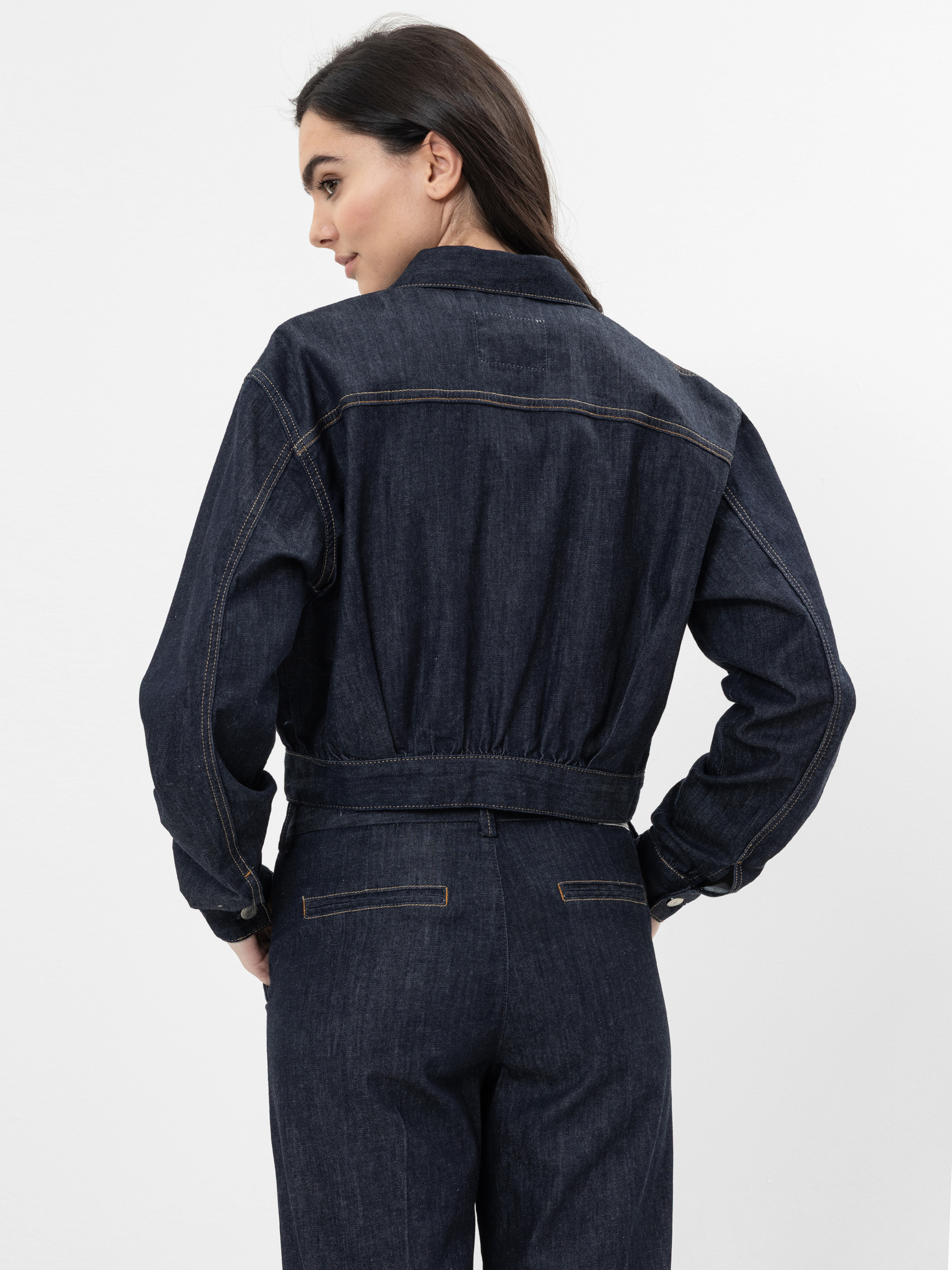Eine Frau mit langen dunklen Haaren steht seitlich und schaut über ihre Schulter. Sie trägt eine dunkle Jeansjacke und eine passende Hose. Der Hintergrund ist einfarbig weiß.