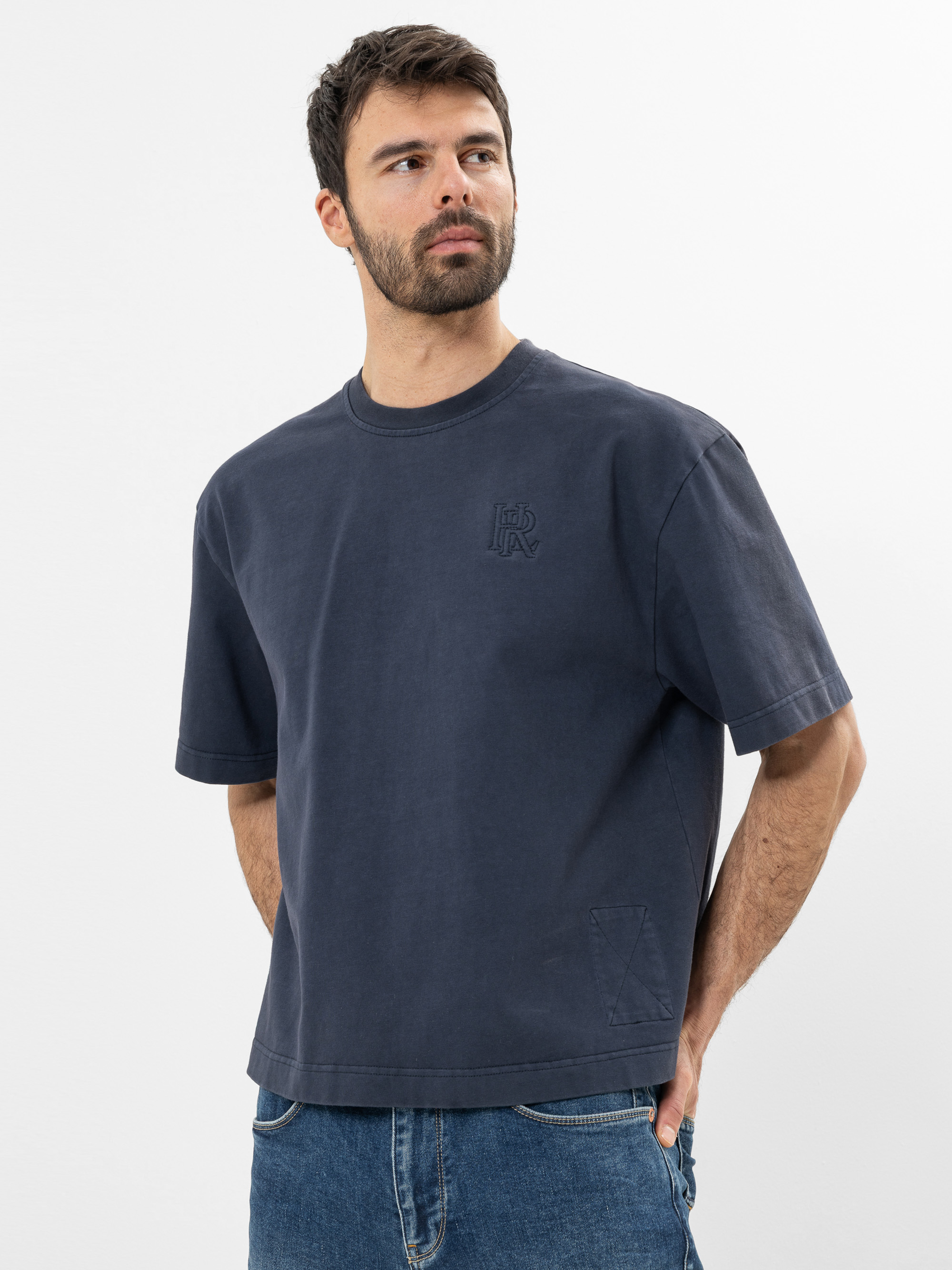 Ein Mann mit kurzen dunklen Haaren und einem Bart trägt ein locker sitzendes dunkelblaues T-Shirt und blaue Jeans. Er steht vor einem schlichten weißen Hintergrund, hat die Hände in den Taschen und schaut leicht zur Seite.