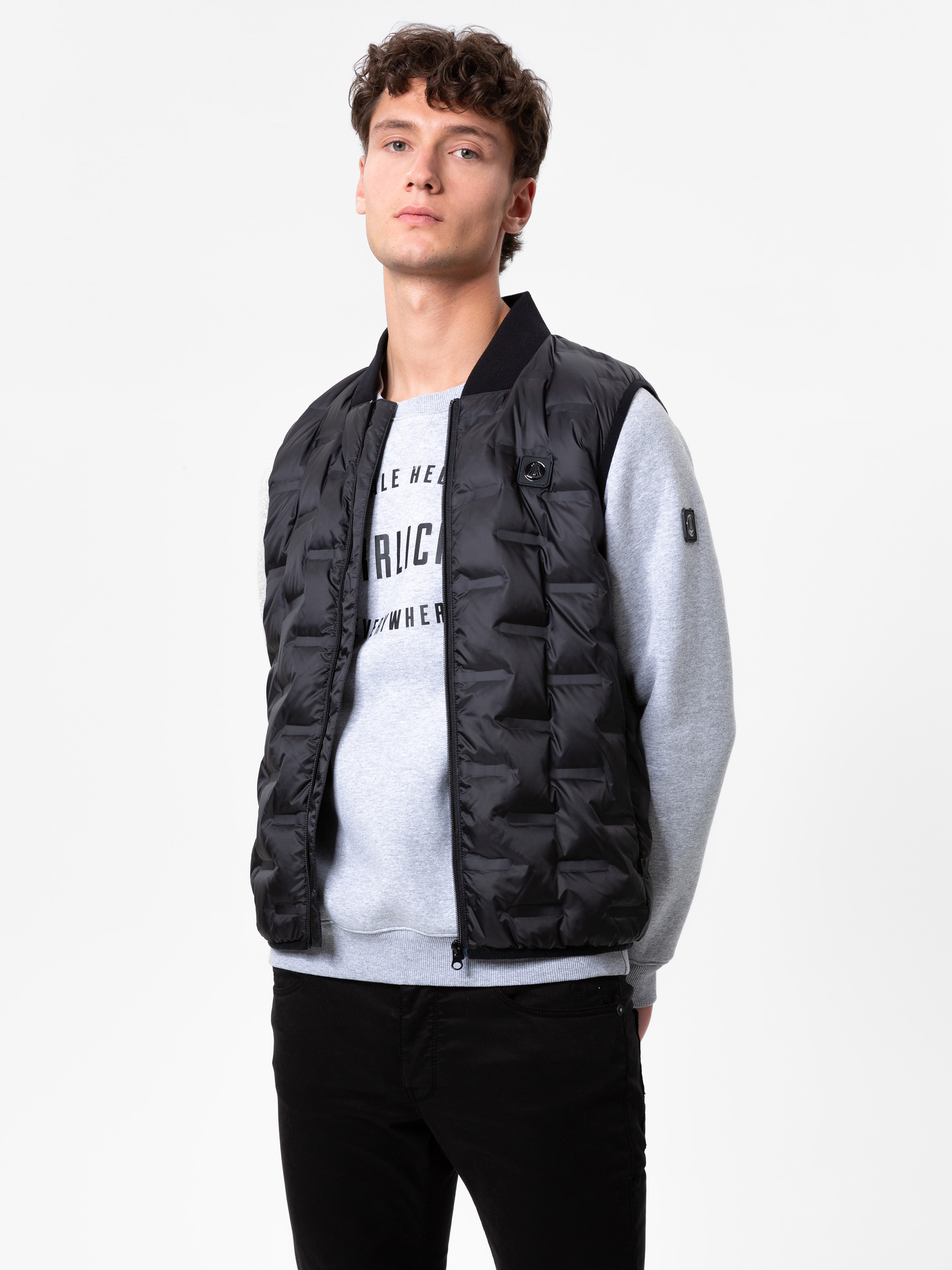 Ein junger Mann mit kurzen braunen Haaren trägt eine schwarze Steppweste über einem grauen Sweatshirt mit Text und eine schwarze Hose. Er steht vor einem schlichten weißen Hintergrund und blickt mit neutraler Miene leicht nach oben.