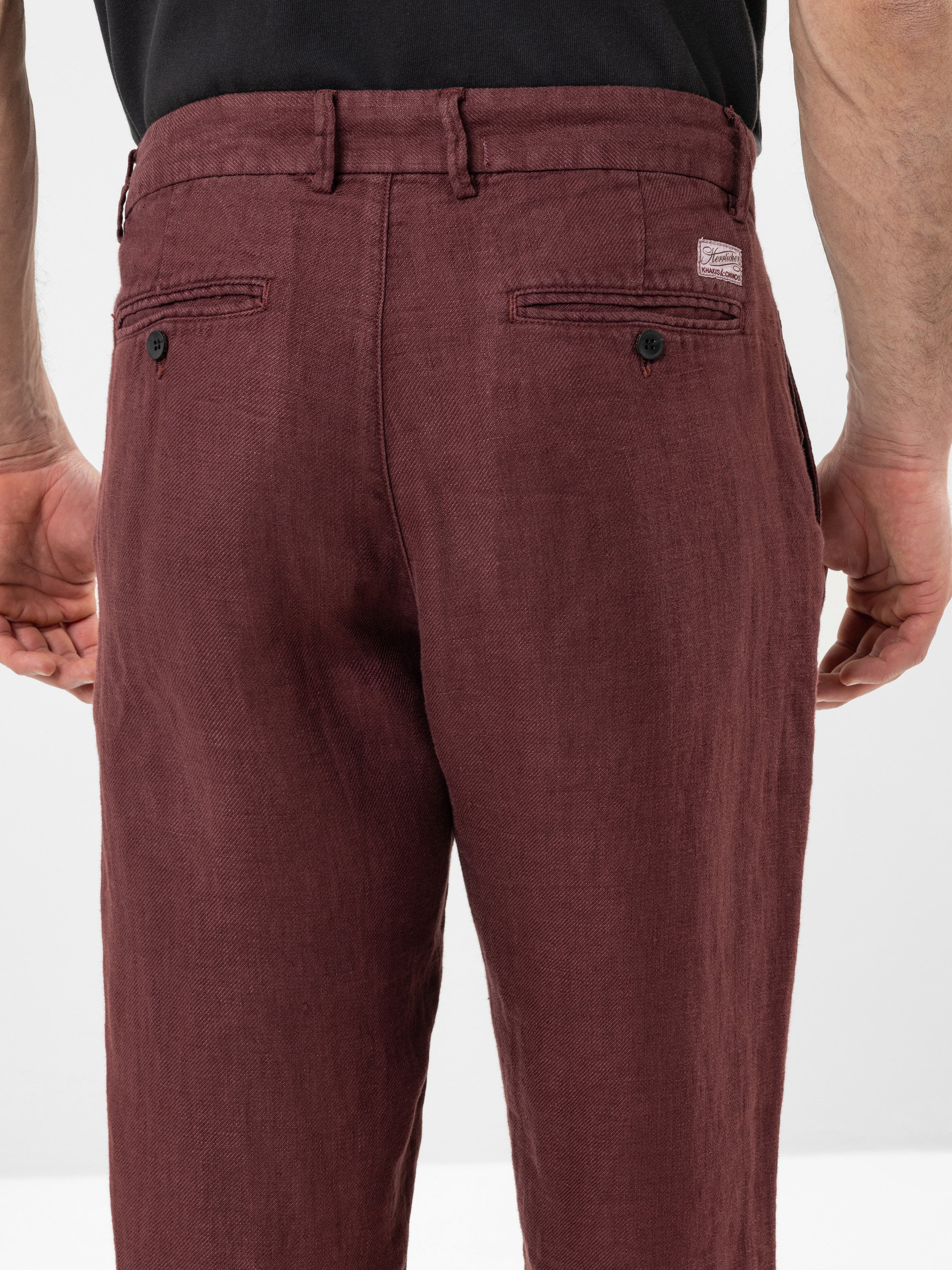 Eine Person, die eine kastanienbraune Leinenhose und ein schwarzes Hemd trägt, wird von hinten gezeigt. Die Hose hat zwei Gesäßtaschen mit Knöpfen und Gürtelschlaufen. Die Arme der Person liegen entspannt an der Seite.