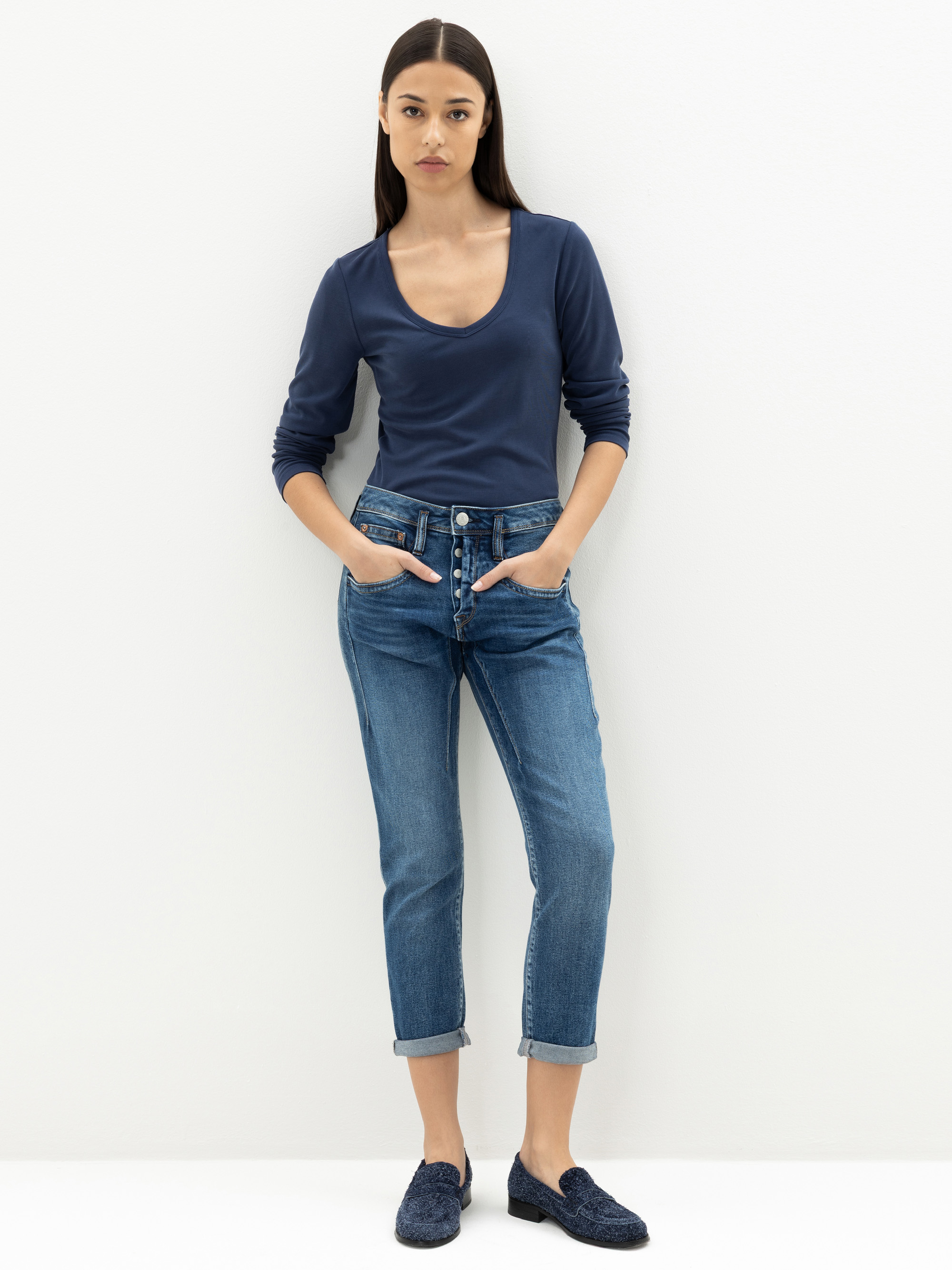 Eine Frau steht vor einer schlichten weißen Wand und trägt ein marineblaues langärmeliges Oberteil, blaue Jeans mit Knöpfen und marineblaue Halbschuhe. Ihre Hände stecken in den Taschen und sie hat glattes, dunkles Haar.