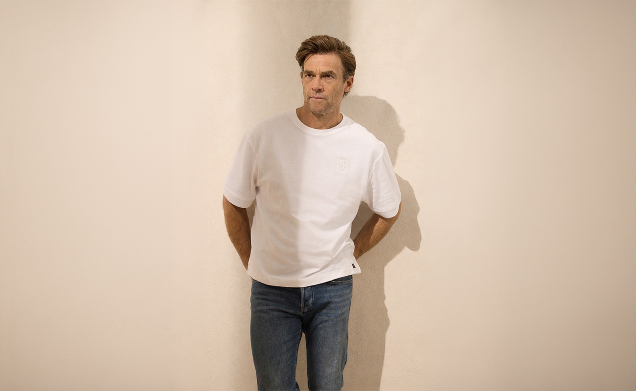 Ein Mann in weißem T-Shirt und blauer Jeans steht mit den Händen auf dem Rücken an einer beigen Wand und schaut leicht zur Seite. Sein Schatten fällt auf die Wand hinter ihm.