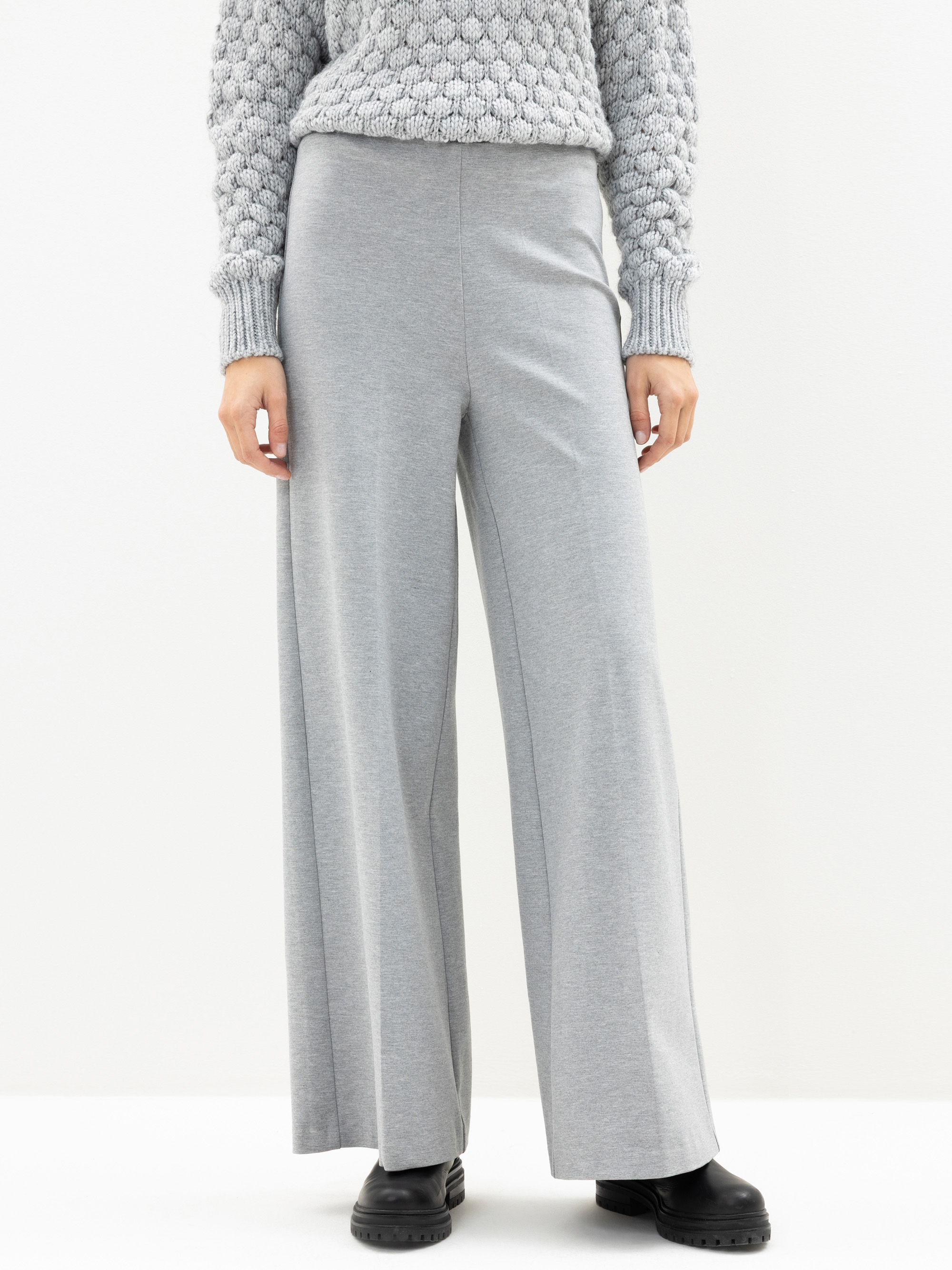 Eine Person, die eine hellgraue Hose mit weitem Bein, einen hellgrauen Pullover mit Struktur und schwarze Schuhe trägt, steht vor einem schlichten weißen Hintergrund. Das Gesicht der Person ist nicht sichtbar.