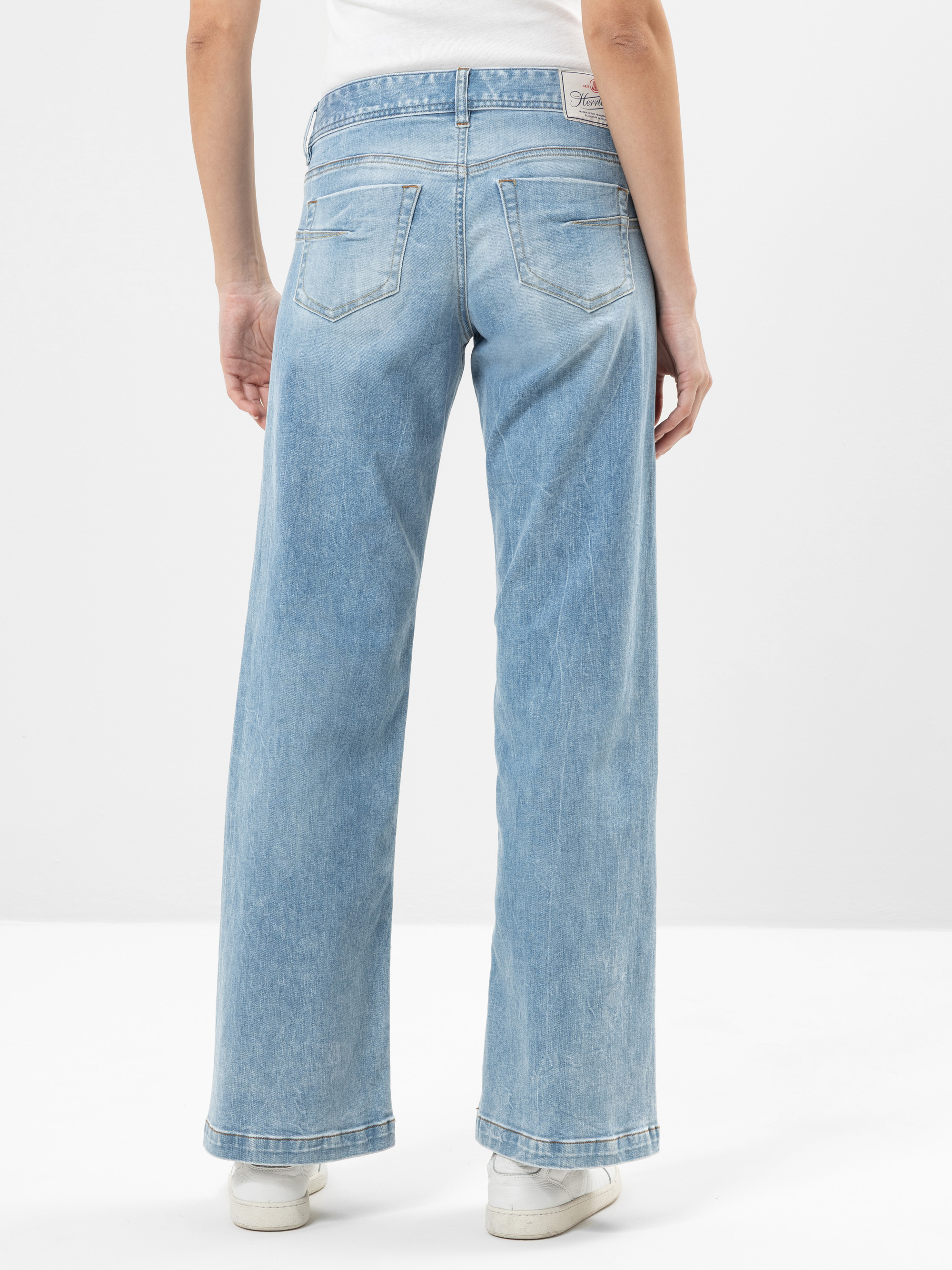 Eine Person, die eine hellblaue, hochgekrempelte Jeans mit weitem Bein und weiße Turnschuhe trägt, steht mit dem Gesicht nach außen vor einem schlichten weißen Hintergrund.
