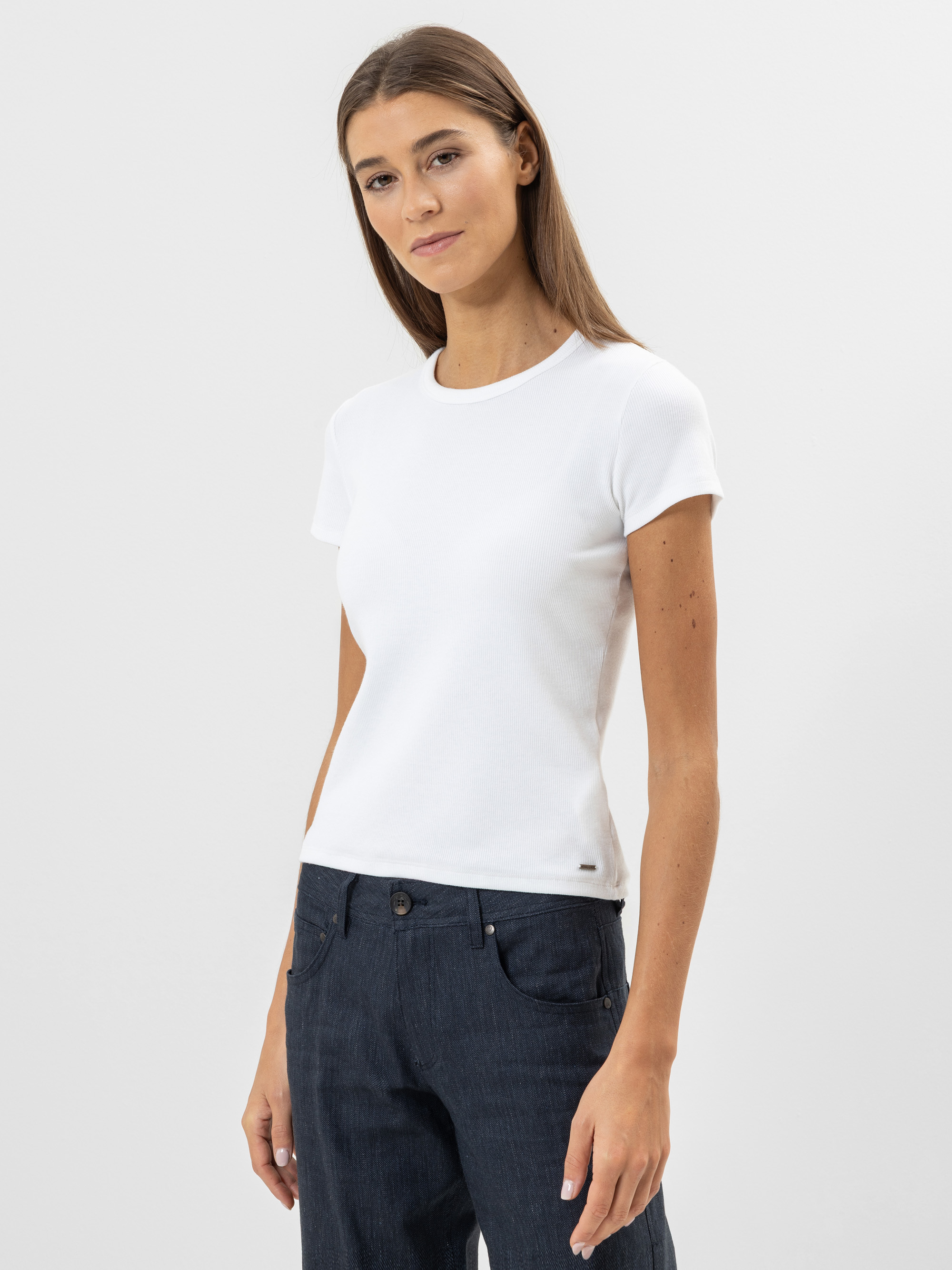 Eine Frau mit glattem braunem Haar, die ein weißes, kurzärmeliges T-Shirt und dunkelblaue Jeans trägt, steht vor einem schlichten hellgrauen Hintergrund und blickt mit neutralem Blick leicht zur Seite.