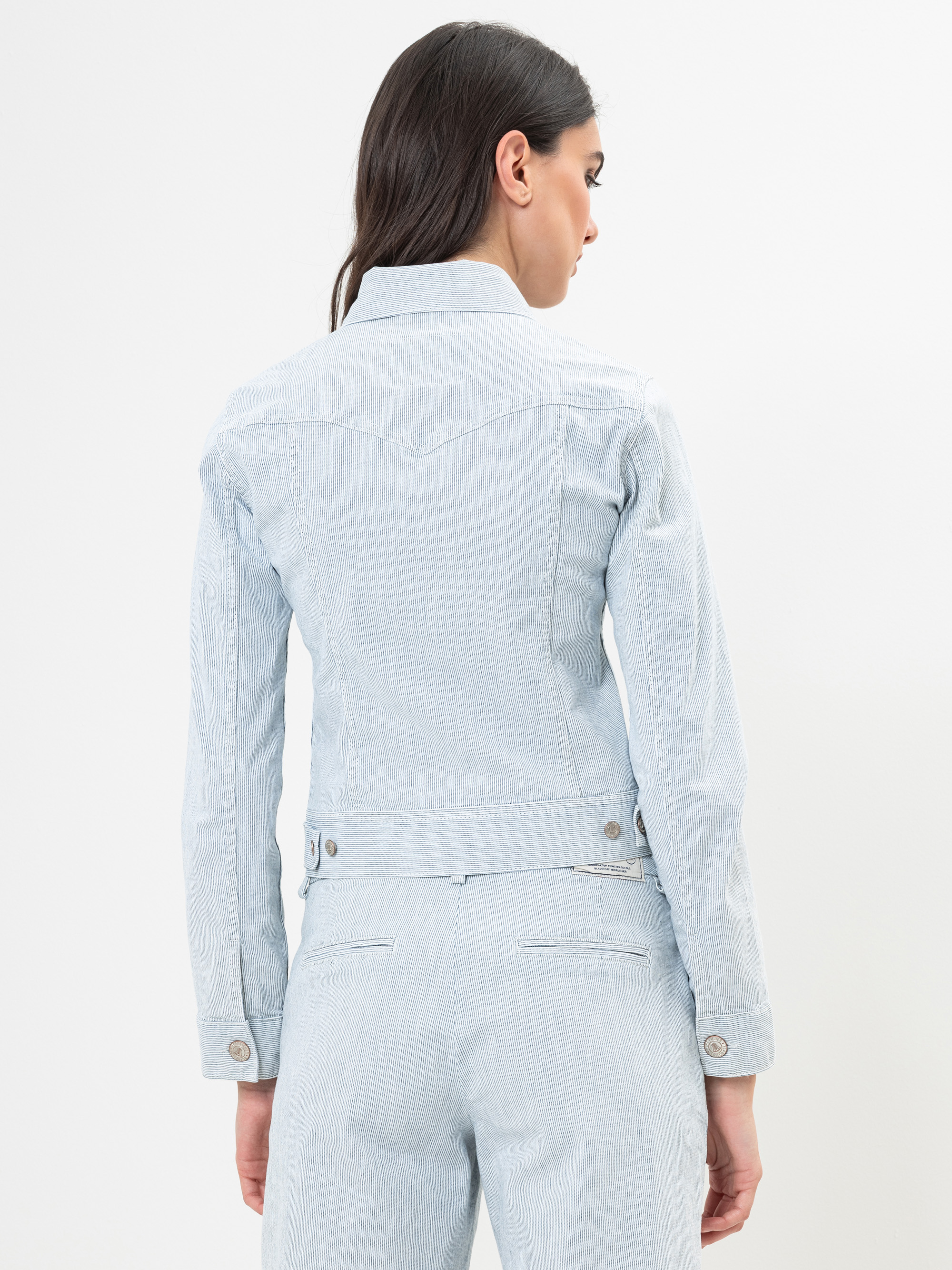 Eine Frau mit langen dunklen Haaren, die eine hellblaue Jeansjacke und dazu passende Jeans trägt, steht mit dem Gesicht zur Kamera vor einem schlichten weißen Hintergrund.