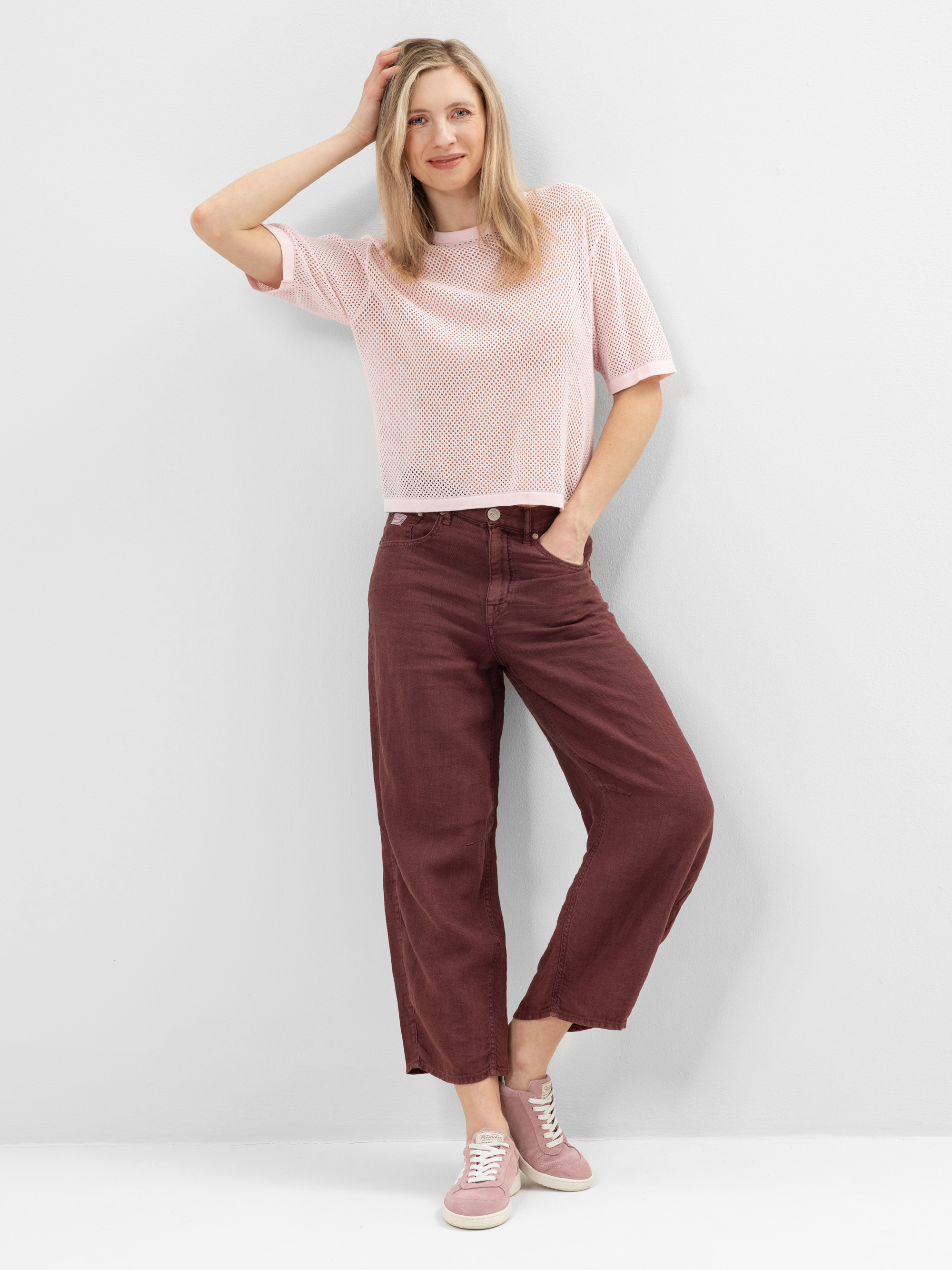 Eine Frau steht lächelnd an einer weißen Wand und stützt sich mit einer Hand auf ihren Kopf. Sie trägt ein blassrosa kurzärmeliges Oberteil, eine weite kastanienbraune Hose und rosa Turnschuhe, eine Hand in der Tasche.