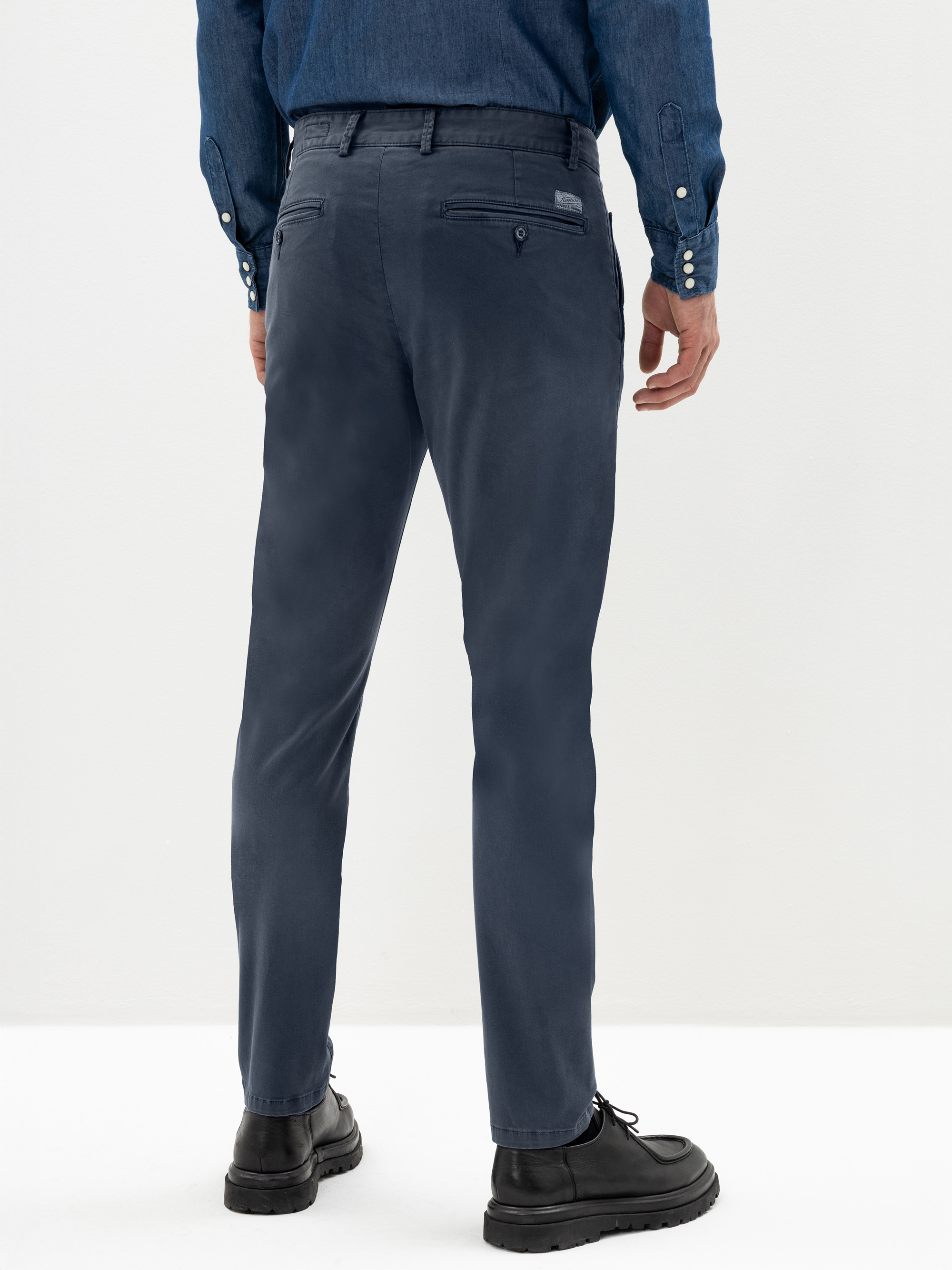 Eine Person, die eine dunkelblaue Hose, ein Jeanshemd und schwarze Schuhe trägt, steht von der Kamera abgewandt vor einem schlichten weißen Hintergrund.