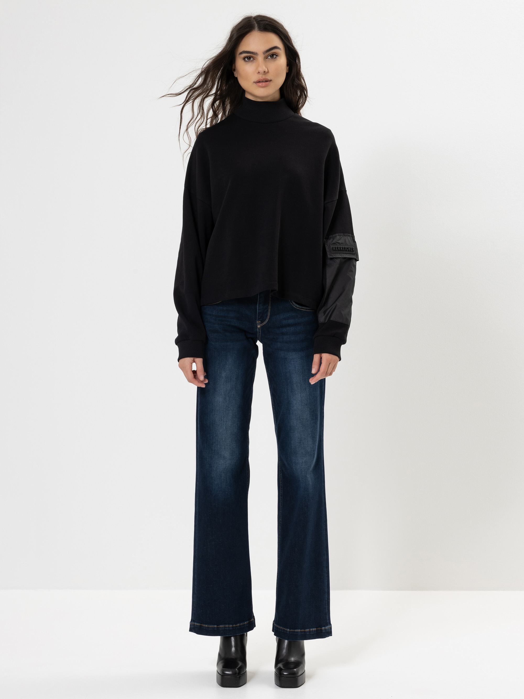 Eine Frau steht vor einem schlichten weißen Hintergrund und trägt einen weiten schwarzen Rollkragenpullover, dunkelblaue Jeans mit weitem Bein und schwarze Schuhe mit Absätzen. Sie hat langes gewelltes Haar und blickt mit neutralem Blick in die Kamera.