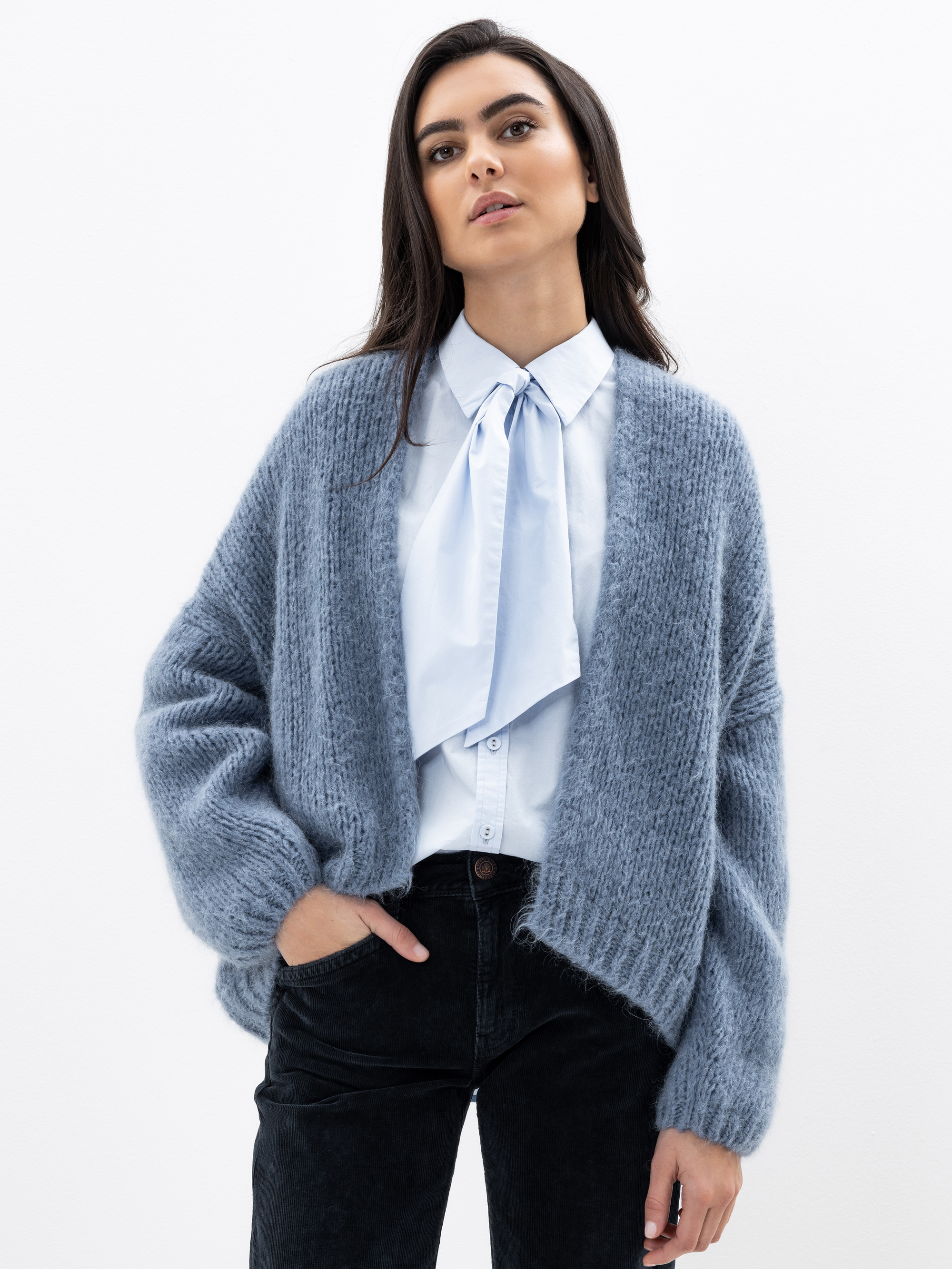 Eine Frau mit langen dunklen Haaren trägt eine hellblaue Bluse mit einer Krawatte, eine dicke graue Strickjacke und eine schwarze Hose. Sie steht vor einem schlichten weißen Hintergrund, hat eine Hand in der Tasche und blickt selbstbewusst in die Kamera