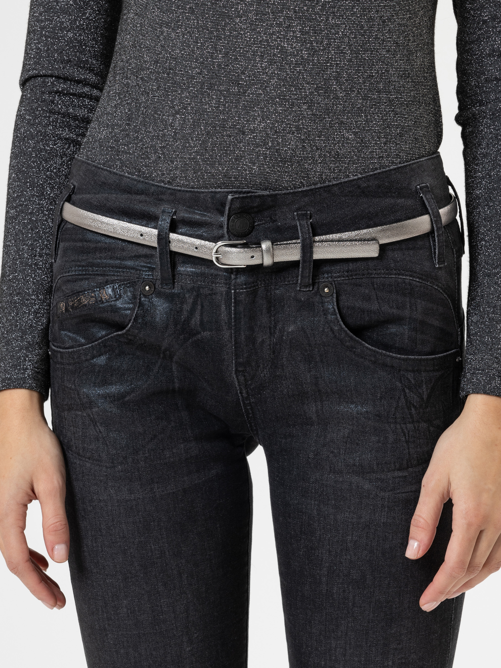 Eine Person, die ein glitzerndes langärmeliges Oberteil, dunkle Jeans und einen silbernen Metallgürtel trägt. Nur der Oberkörper und die Hände sind vor einem einfarbigen Hintergrund zu sehen.