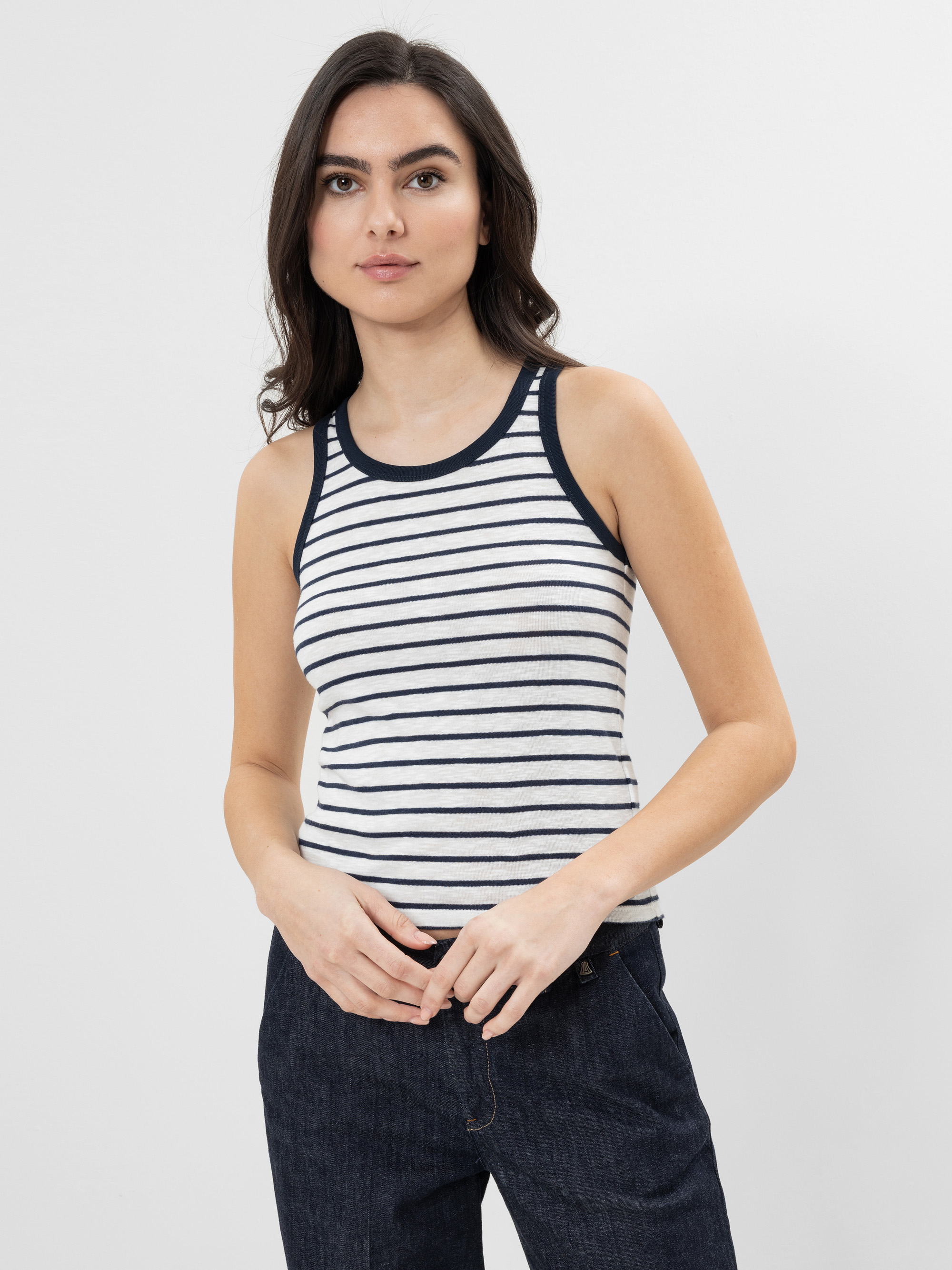 Eine Frau mit langen dunklen Haaren trägt ein ärmelloses, weiß-marinefarben gestreiftes Tanktop und dunkle Jeans und steht vor einem schlichten weißen Hintergrund.