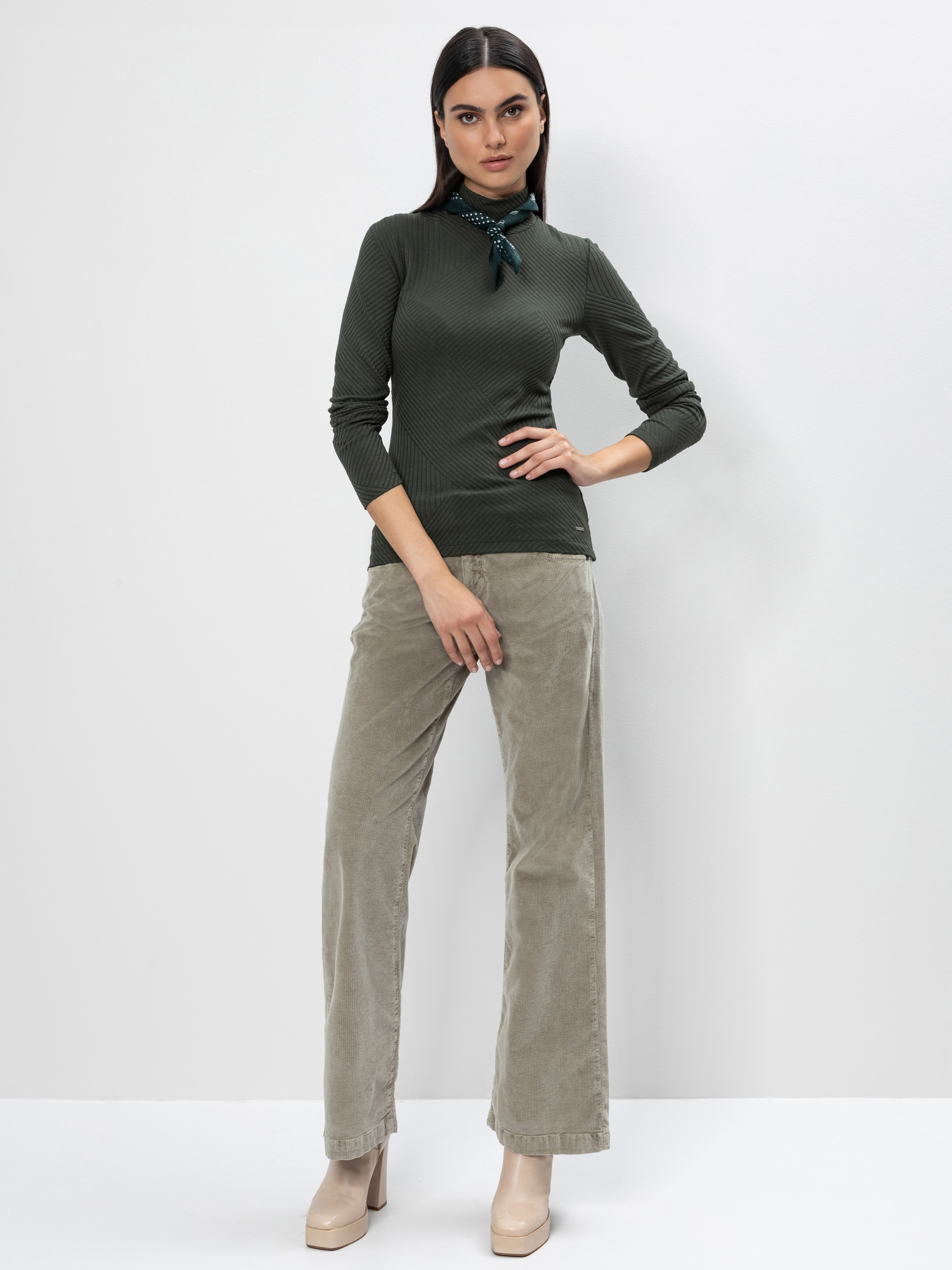 Eine Frau steht vor einem schlichten weißen Hintergrund und trägt einen olivgrünen langärmeligen Pullover, eine hellbeige Cordhose, einen grünen Schal um den Hals und beige Plateauschuhe. Sie posiert mit einer Hand an der Hüfte.