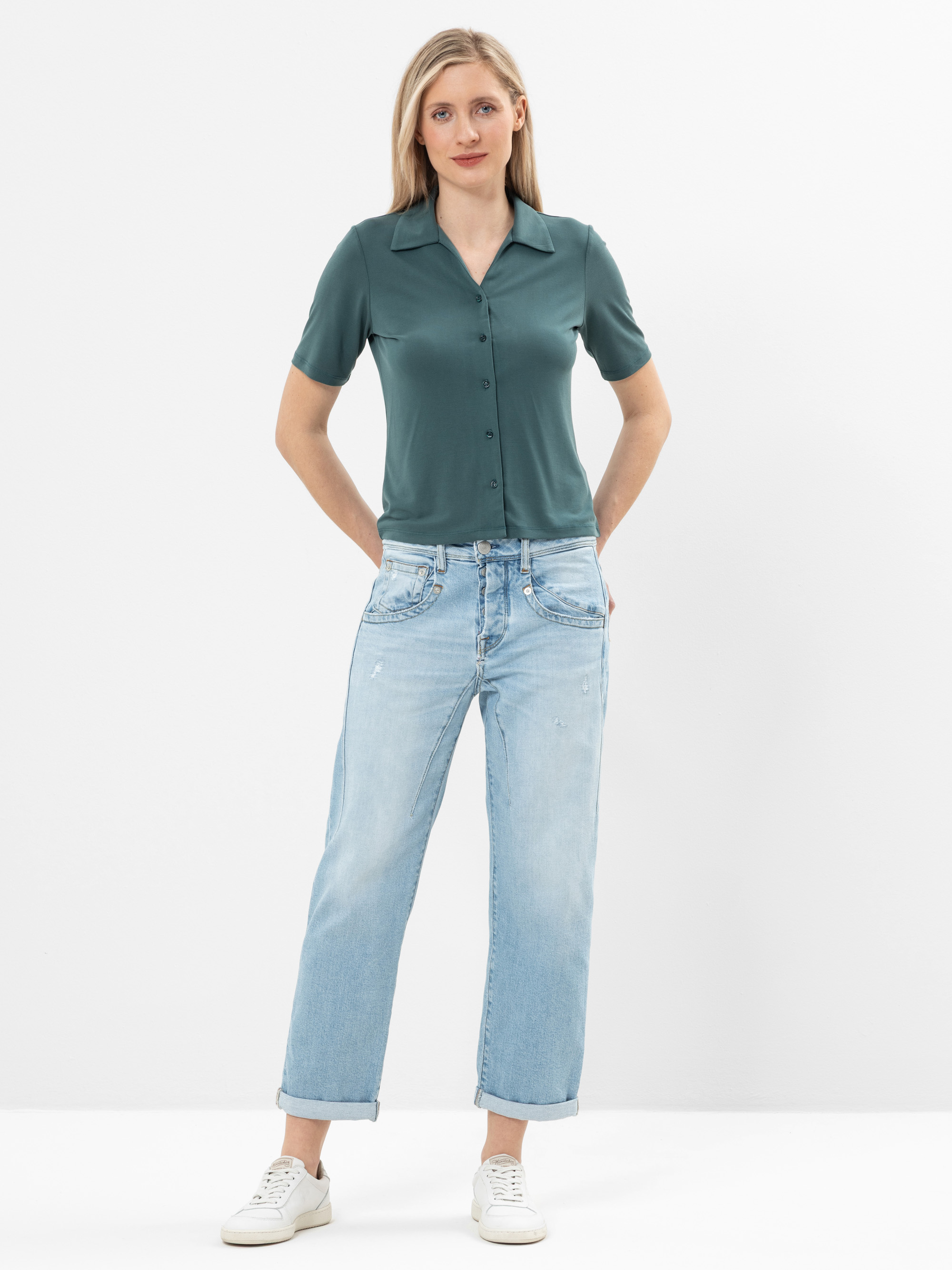 Eine Frau steht vor einem weißen Hintergrund und trägt ein kurzärmeliges, blaugrünes Button-up-Hemd, hellblaue Jeans mit Bündchen und weiße Turnschuhe. Sie hat blonde Haare und ihre Hände stecken in den Gesäßtaschen.