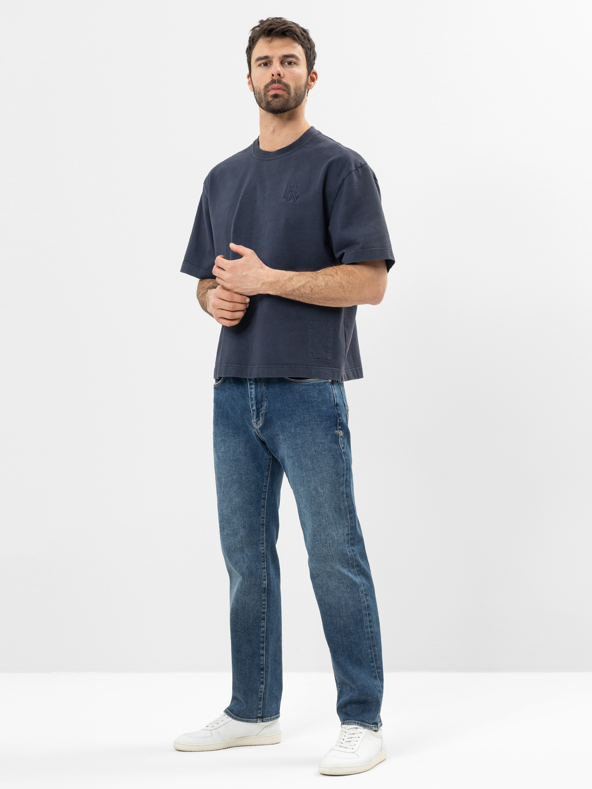 Ein Mann steht in einer Studioumgebung und trägt ein dunkelblaues T-Shirt in Übergröße, blaue Jeans und weiße Turnschuhe. Er hat kurzes dunkles Haar, einen Bart und blickt leicht zur Seite, während er die Hände verschränkt. Der Hintergrund ist ein