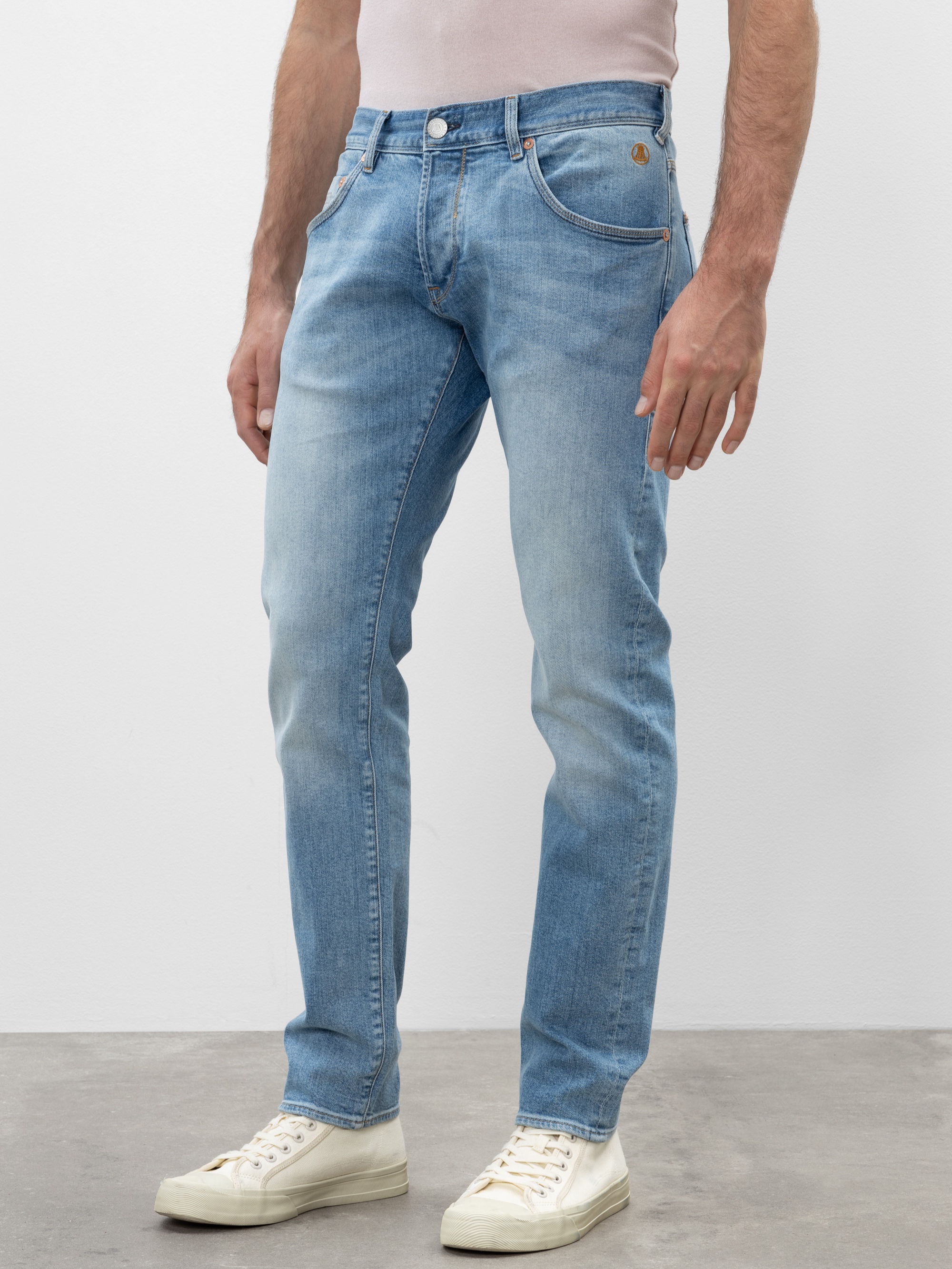 Eine Person steht vor einem schlichten Hintergrund und trägt hellblaue Jeans und weiße Turnschuhe. Die Jeans hat eine klassische Passform mit leichten Ausbleichungen, und die Hände der Person liegen entspannt an der Seite.