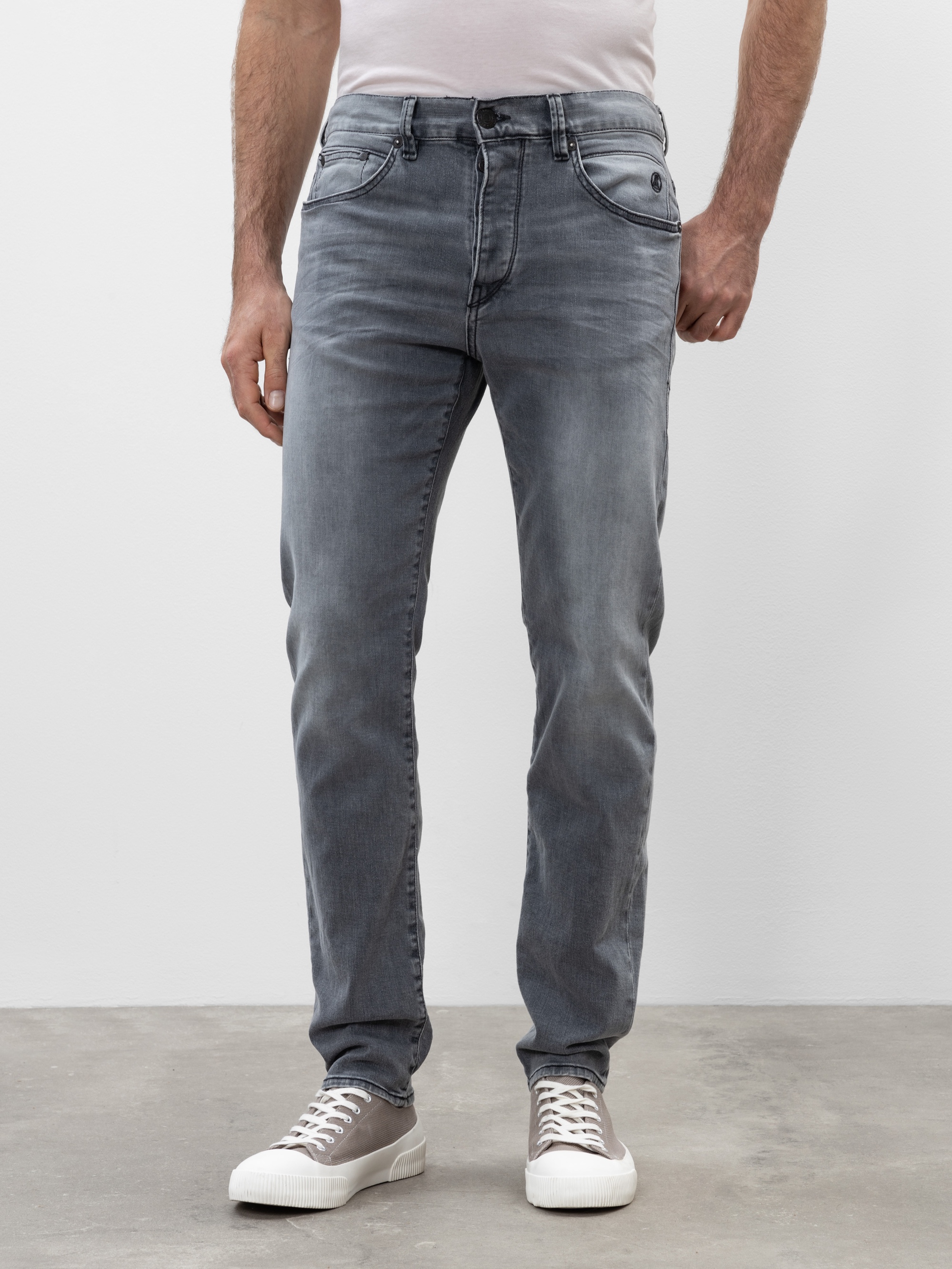 Eine Person, die eine hellgraue Jeans, ein weißes Hemd und graue Turnschuhe trägt, steht auf einem Betonboden vor einem schlichten weißen Hintergrund, wobei nur die untere Hälfte ihres Körpers zu sehen ist.