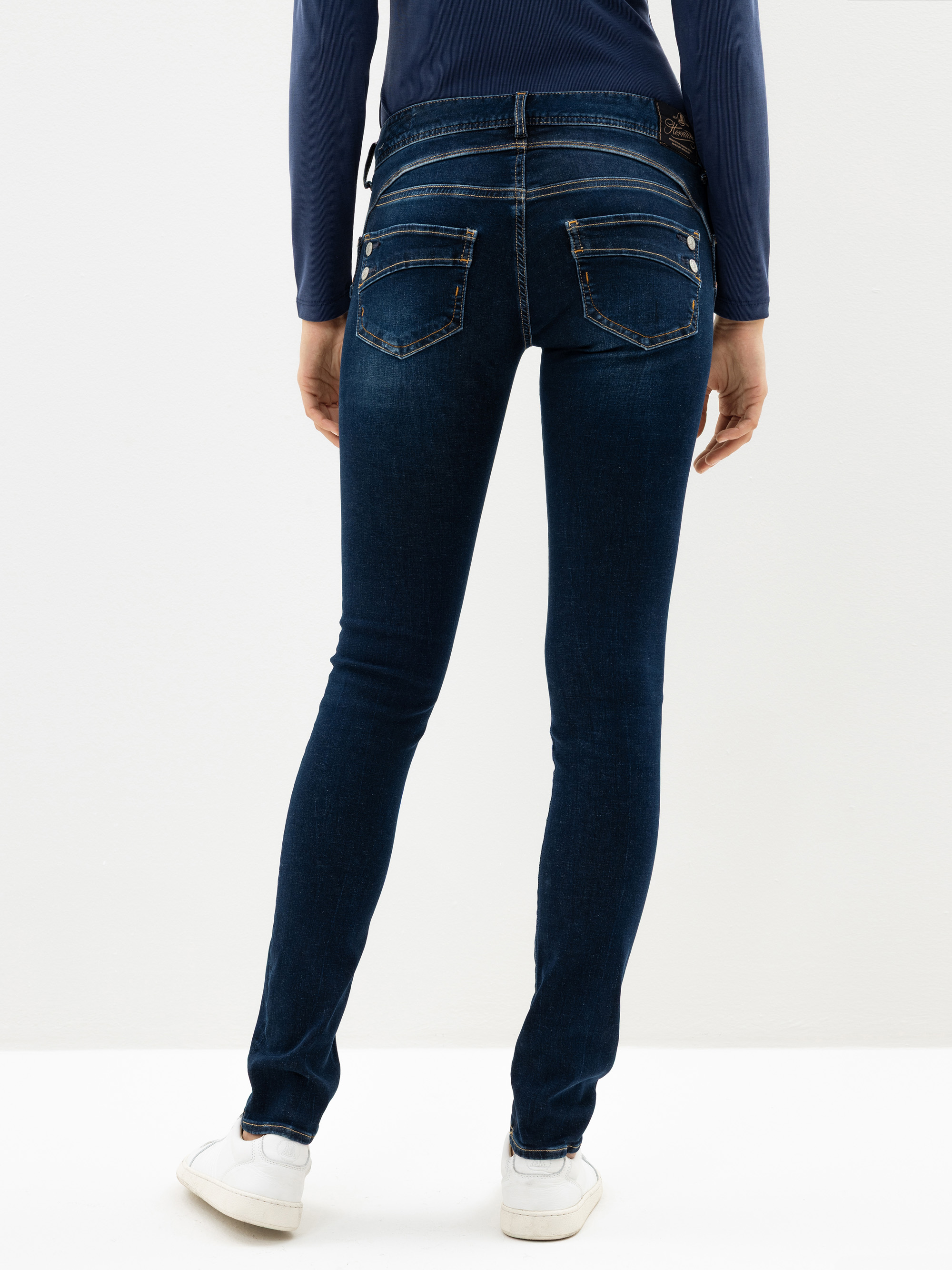 Person, die dunkelblaue Röhrenjeans, ein langärmeliges marineblaues Oberteil und weiße Turnschuhe trägt und mit dem Rücken zur Kamera vor einem schlichten weißen Hintergrund steht.