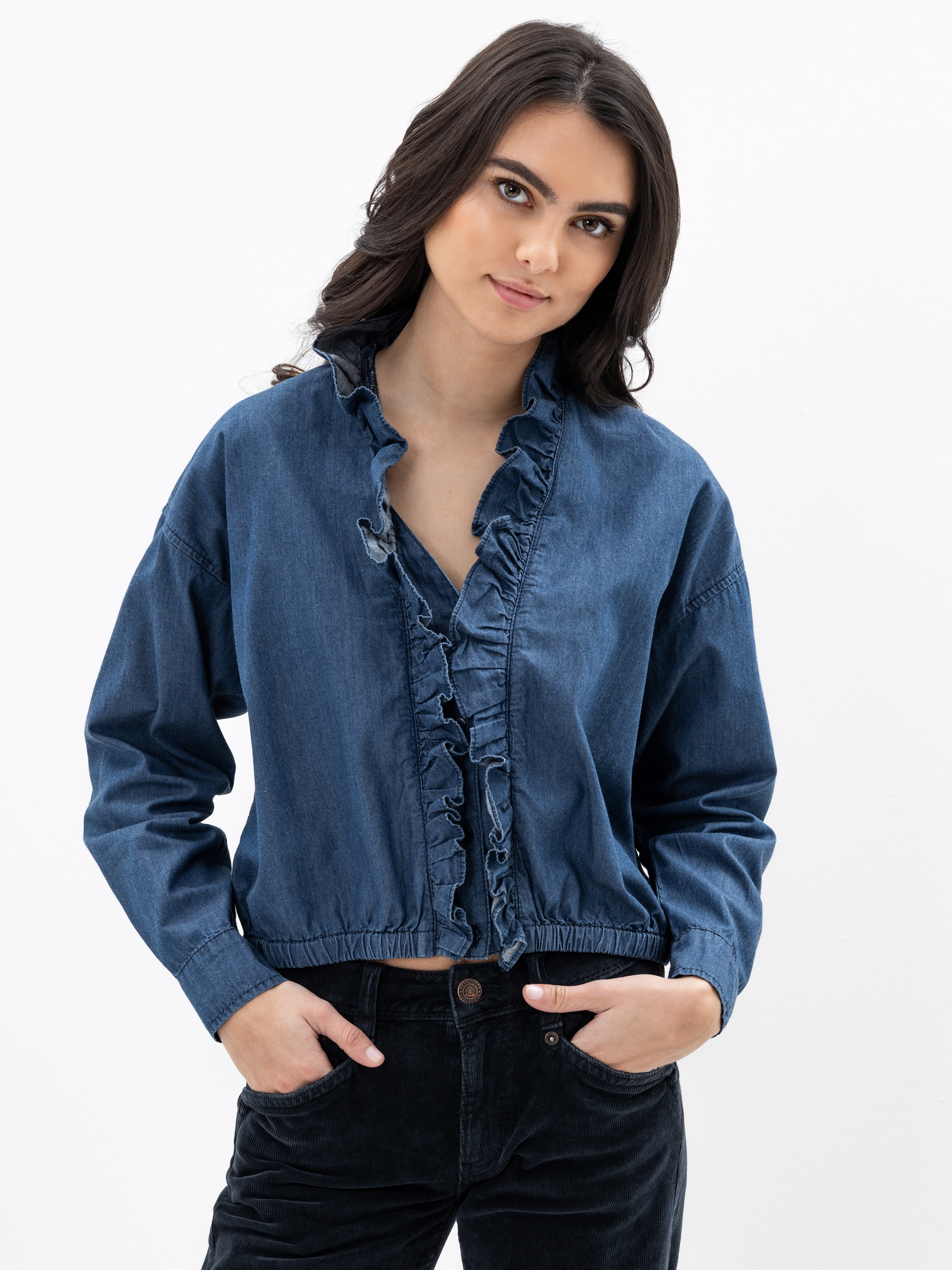 Eine Frau mit langen dunklen Haaren trägt eine Jeansbluse mit Rüschenkragen und eine schwarze Hose. Sie steht vor einem schlichten weißen Hintergrund, hat die Hände in den Taschen und schaut in die Kamera.