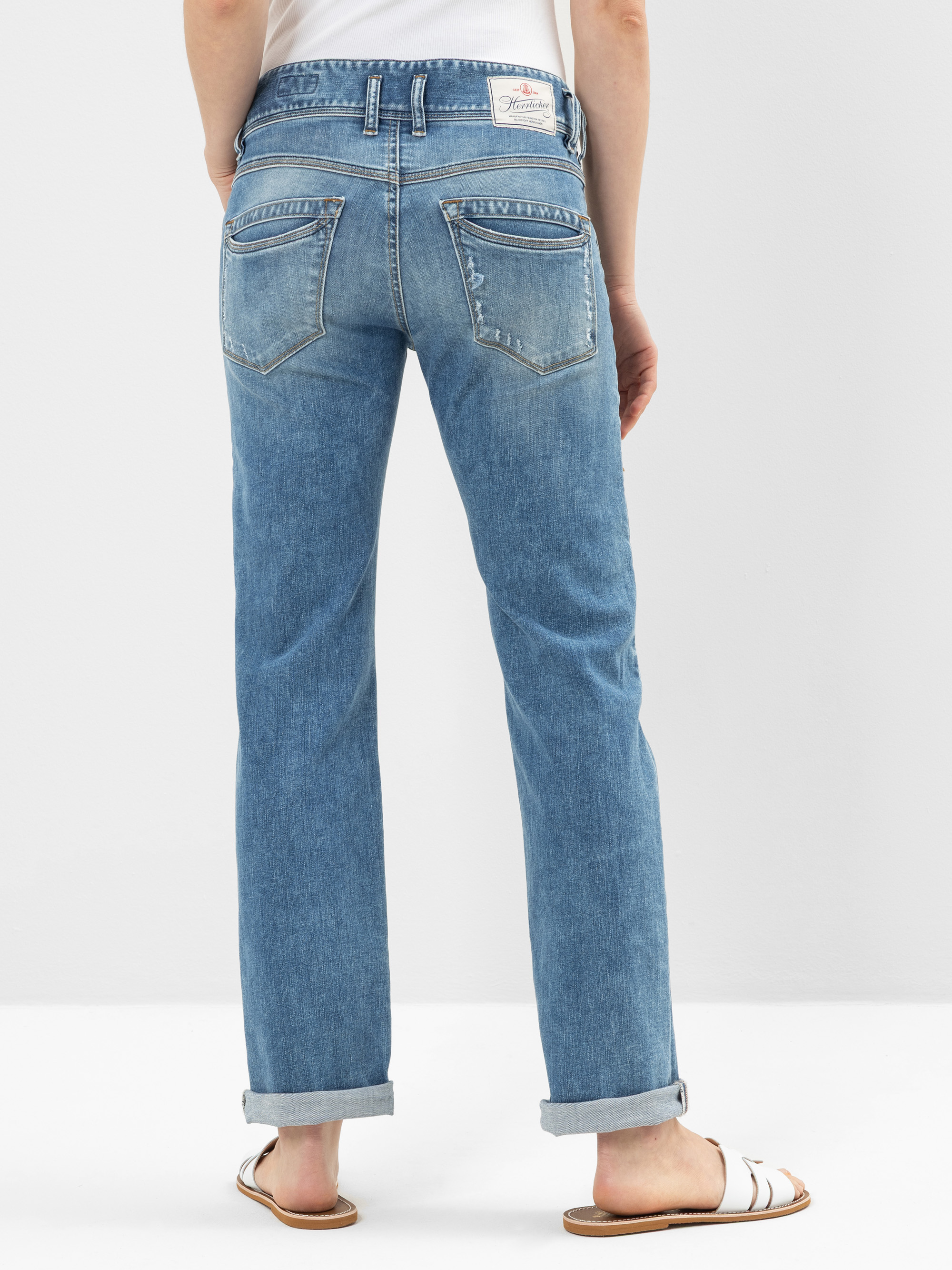 Person, die mit dem Gesicht zur Wand steht und eine hellblaue Jeans mit sichtbaren Gesäßtaschen und einem weißen Aufnäher, ein weißes Oberteil und weiße Sandalen vor einem schlichten hellen Hintergrund trägt.