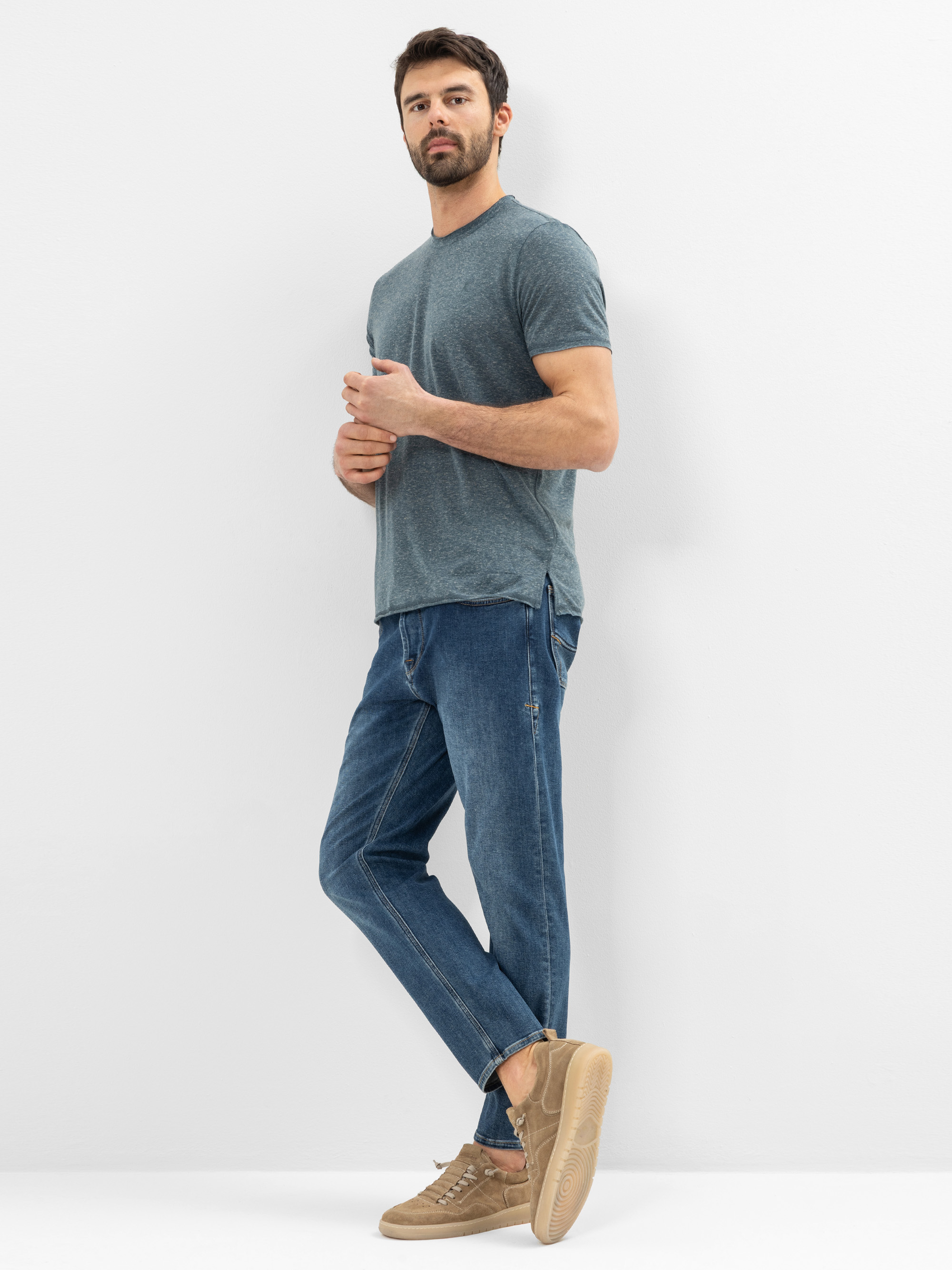 Ein Mann steht vor einer weißen Wand und trägt ein blaugraues T-Shirt, blaue Jeans und hellbraune Turnschuhe. Er hat kurzes dunkles Haar und einen Bart und schaut leicht zur Seite mit einem neutralen Gesichtsausdruck.
