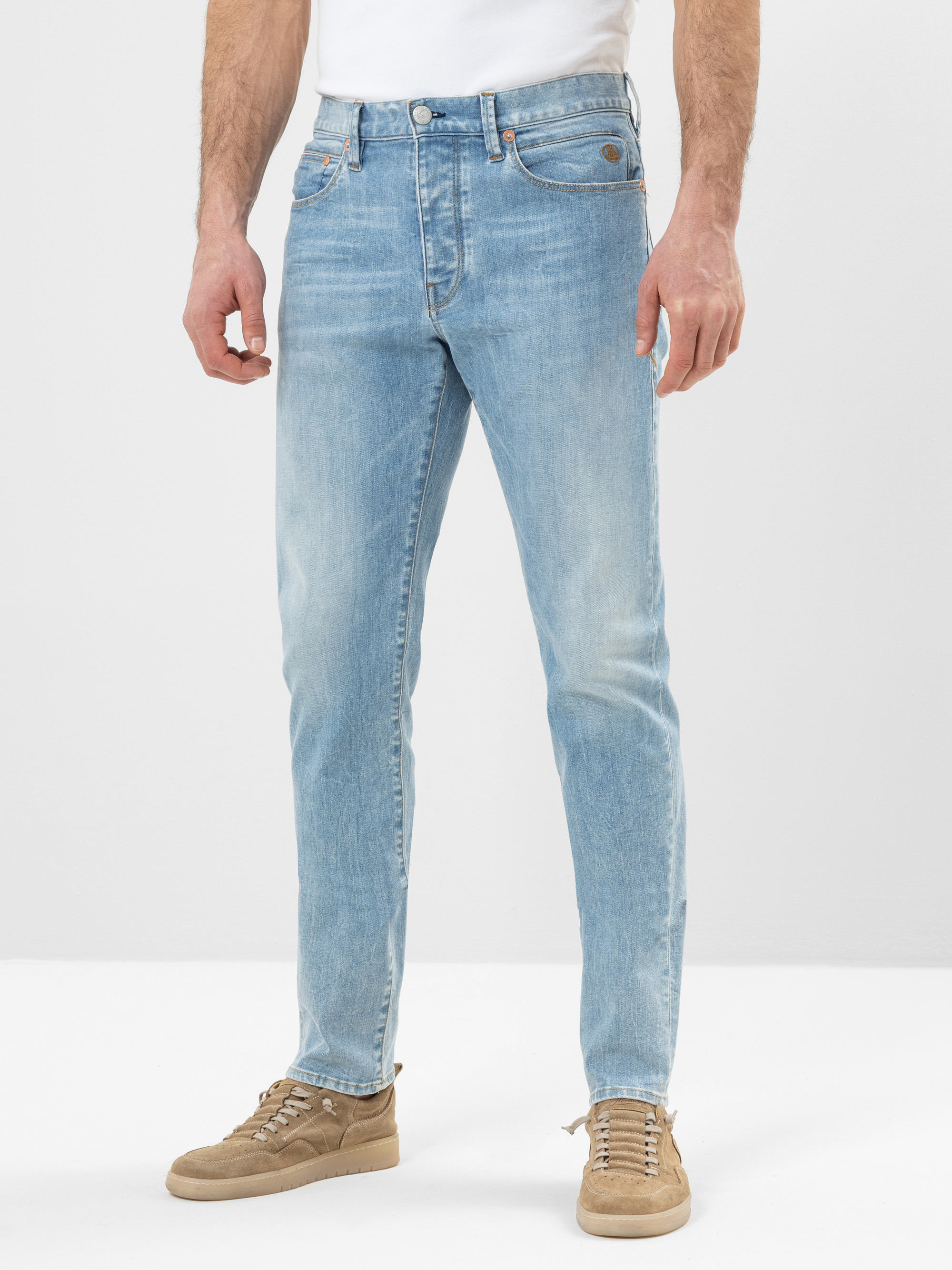 Eine Person, die hellblaue Slim-Fit-Jeans, ein weißes Hemd und beigefarbene Wildlederschuhe trägt, steht vor einem schlichten hellen Hintergrund.