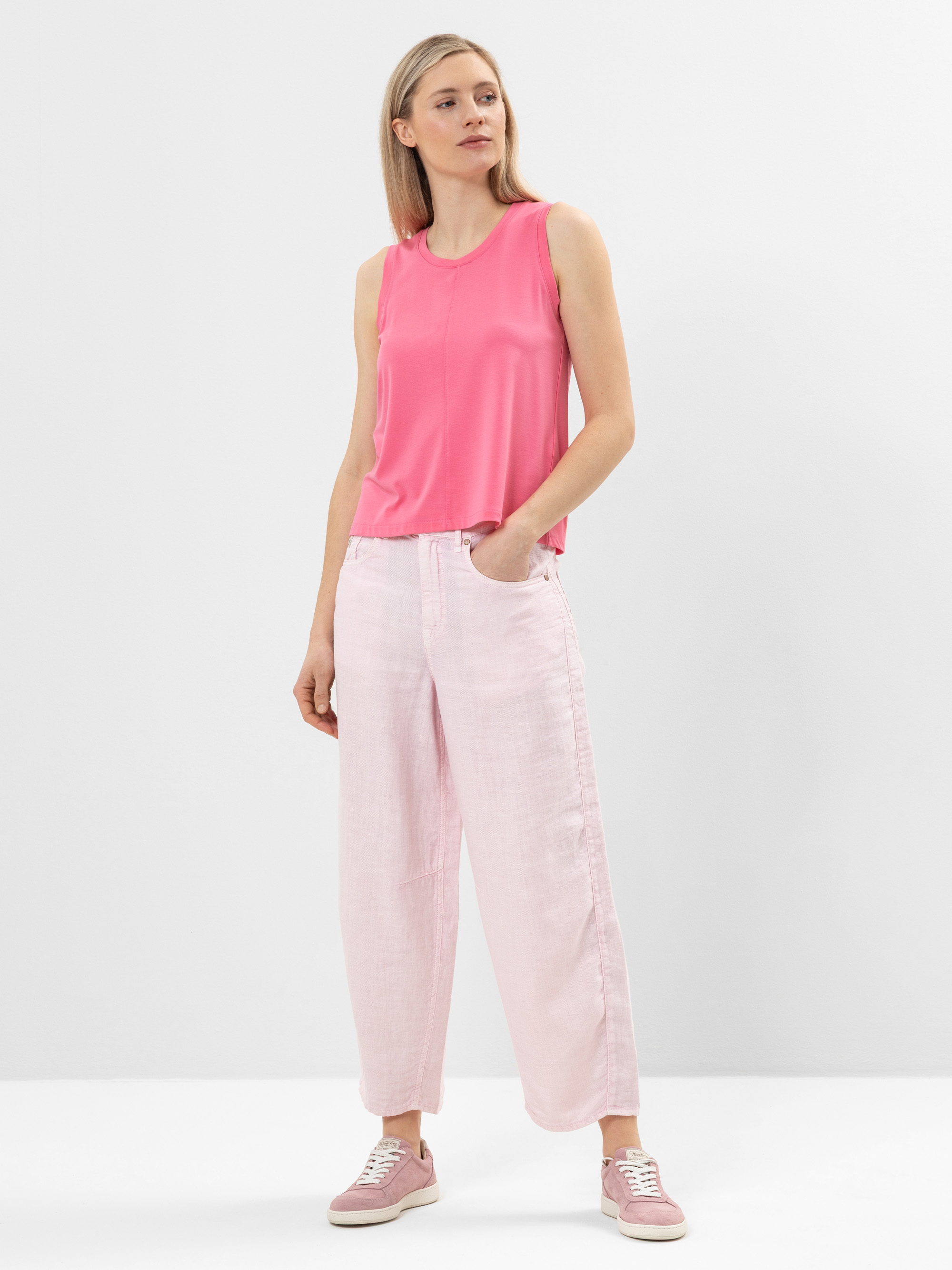 Eine Frau steht vor einem schlichten weißen Hintergrund und trägt ein ärmelloses, hellrosa Oberteil, eine hellrosa Hose mit weitem Bein und dazu passende rosa Turnschuhe. Sie hat helles Haar und ihre rechte Hand steckt in der Hosentasche.