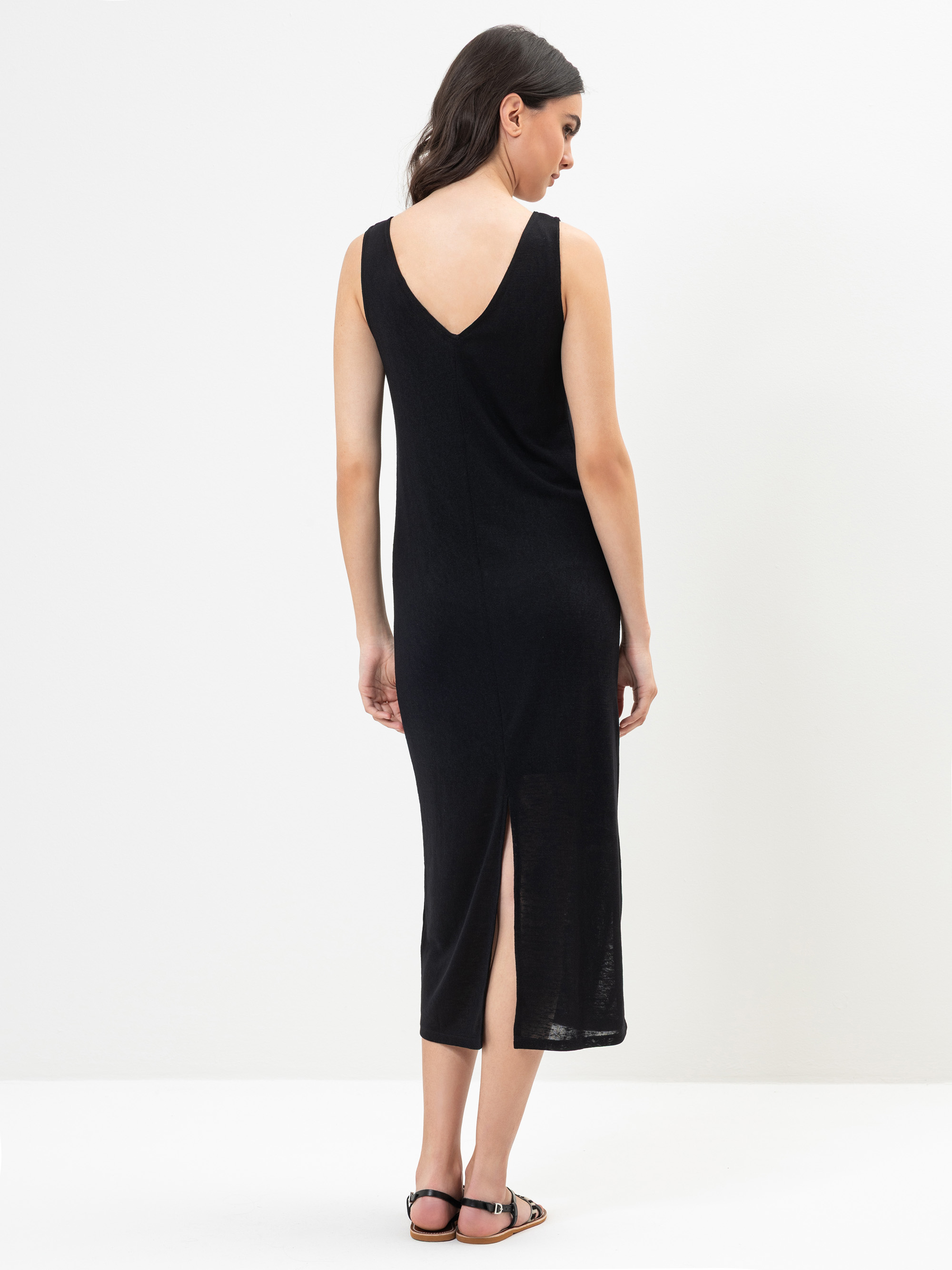 Eine Frau steht mit dem Gesicht zur Wand und trägt ein ärmelloses, schwarzes Midikleid mit V-Ausschnitt und Rückenschlitz. Sie hat langes dunkles Haar und trägt flache Sandalen vor einem schlichten weißen Hintergrund.