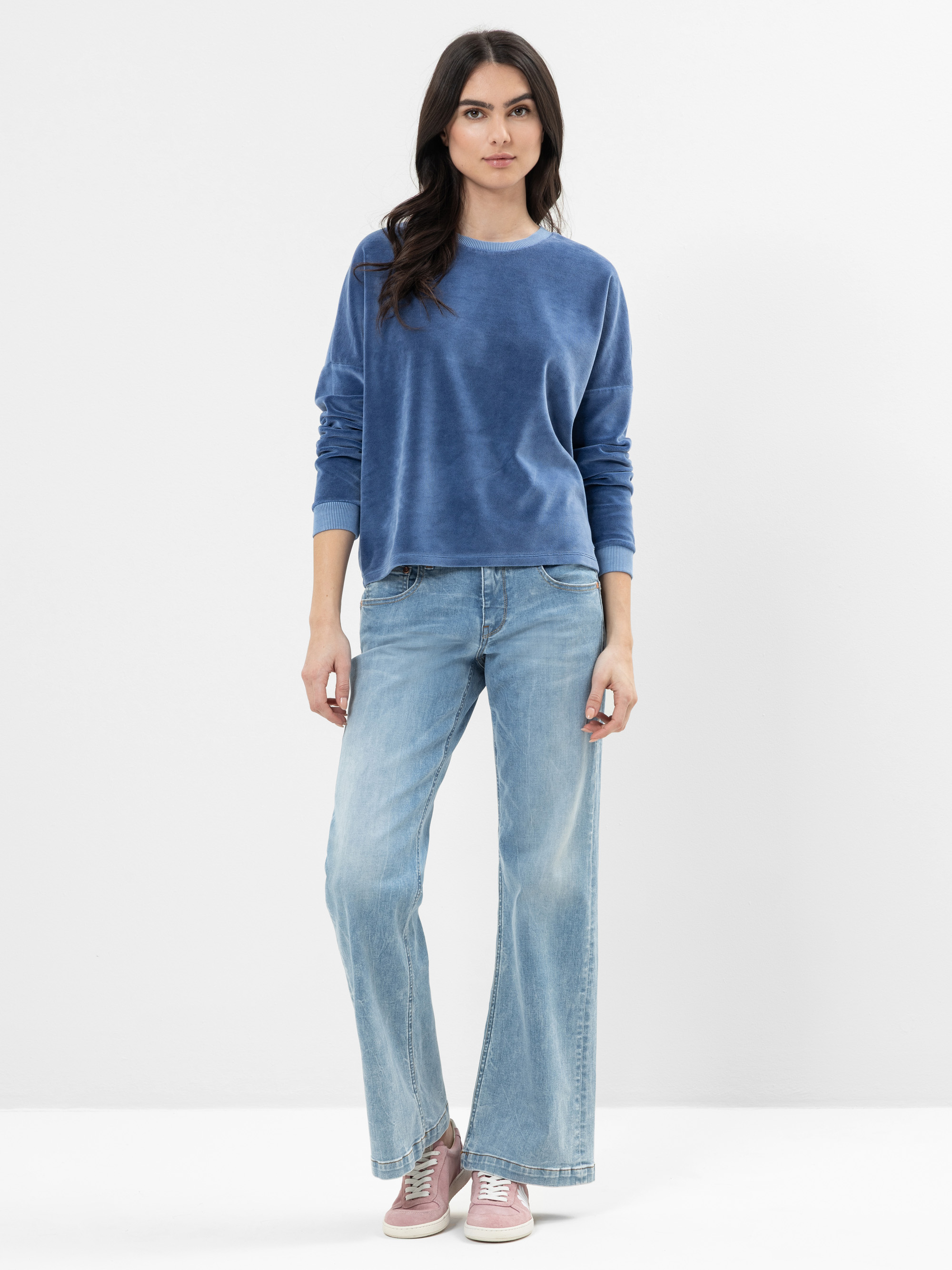 Eine Frau mit langen dunklen Haaren, die ein blaues Samtsweatshirt, hellblaue Jeans mit weitem Bein und rosa Turnschuhe trägt, steht vor einem schlichten weißen Hintergrund.