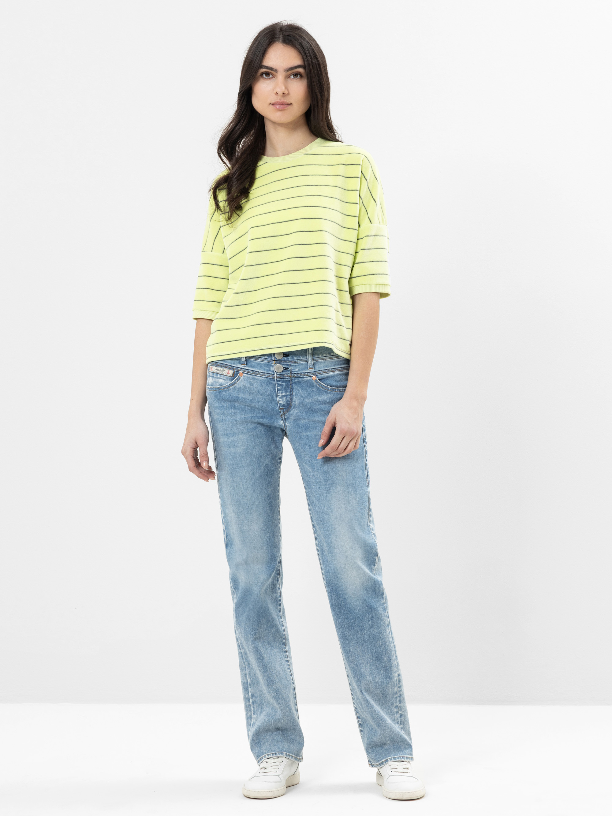 Eine Frau steht vor einem schlichten weißen Hintergrund und trägt ein gelbes, kurzärmeliges, gestreiftes Oberteil, hellblaue Jeans und weiße Turnschuhe. Sie hat langes, dunkles Haar und einen neutralen Gesichtsausdruck.