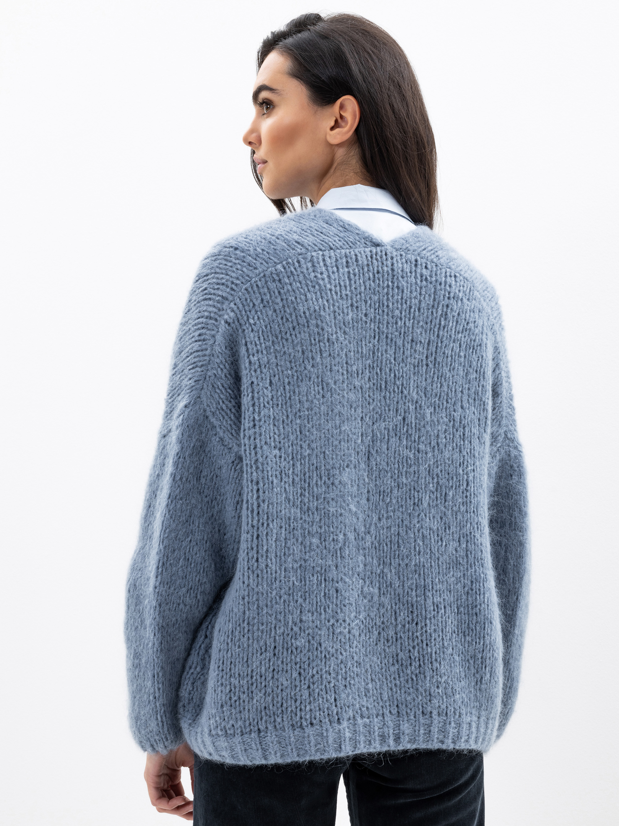 Eine Frau mit dunklem, glattem Haar wird von hinten gezeigt. Sie trägt einen gemütlichen, übergroßen blauen Strickpullover über einem weißen Hemd mit Kragen und einer schwarzen Hose und steht vor einem schlichten weißen Hintergrund.