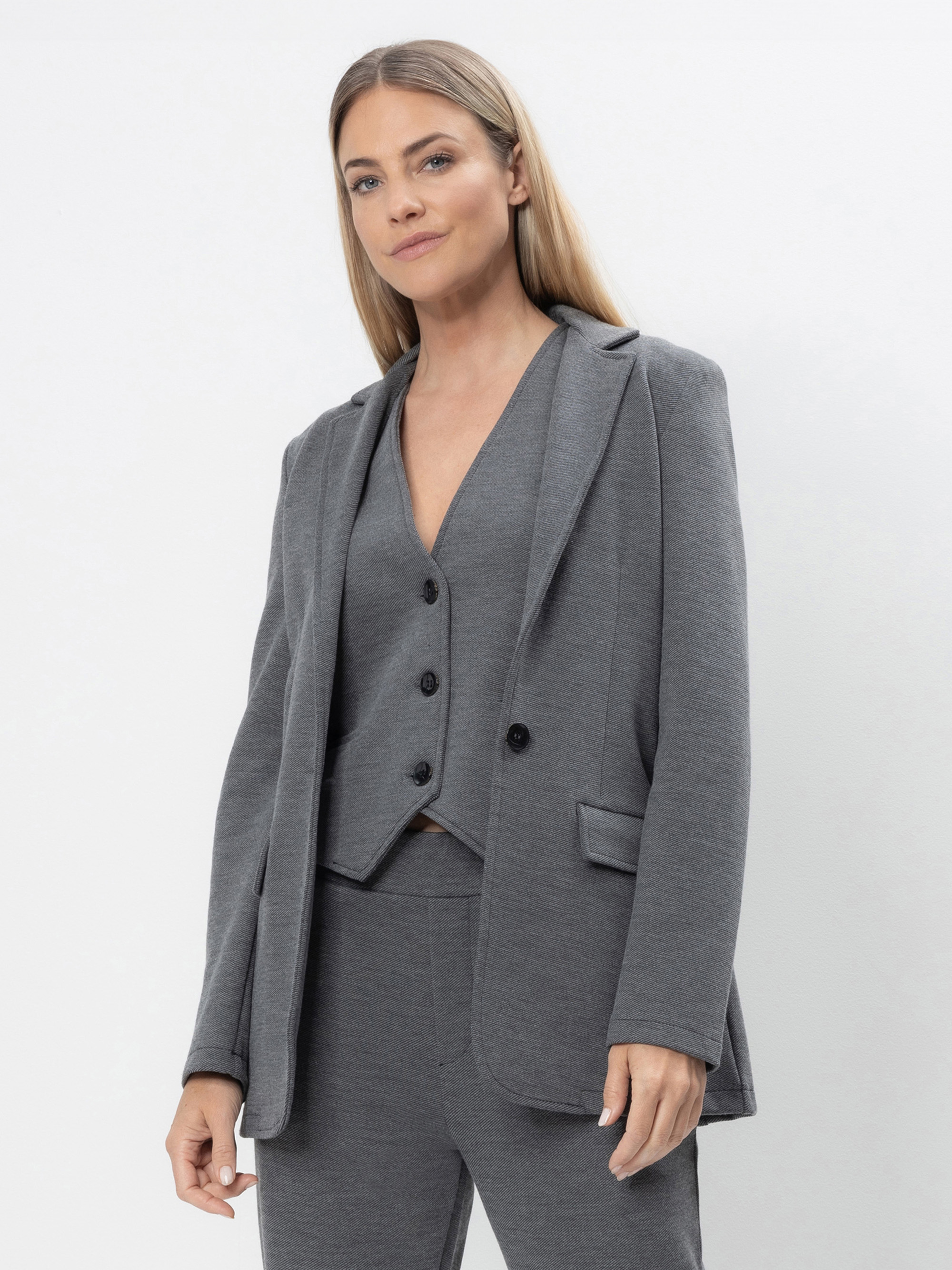 Eine Frau mit langem, glattem blondem Haar trägt einen grauen Anzug mit passender Weste und Blazer, steht vor einem schlichten weißen Hintergrund und blickt selbstbewusst in die Kamera.