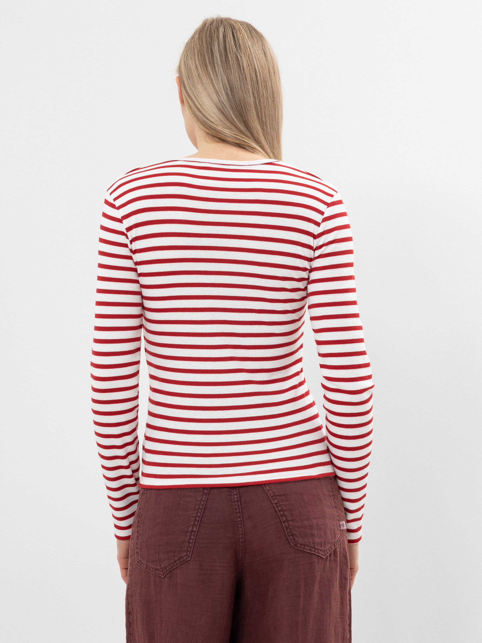 Eine Person mit langen blonden Haaren ist von hinten zu sehen. Sie trägt ein weißes langärmeliges Oberteil mit roten Querstreifen und eine kastanienbraune Hose mit hoher Taille vor einem schlichten weißen Hintergrund.