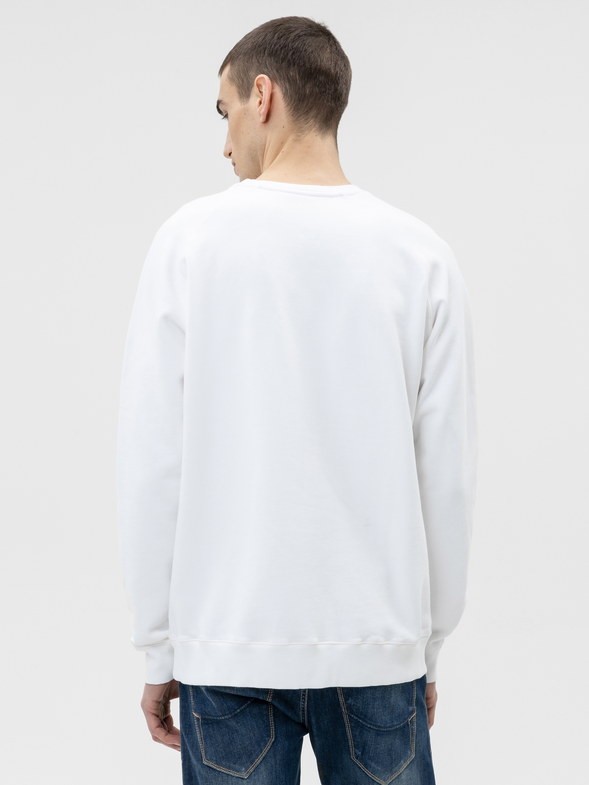 Ein Mann mit kurzen braunen Haaren wird von hinten gezeigt. Er trägt ein schlichtes weißes Sweatshirt und blaue Jeans vor einem hellgrauen Hintergrund.