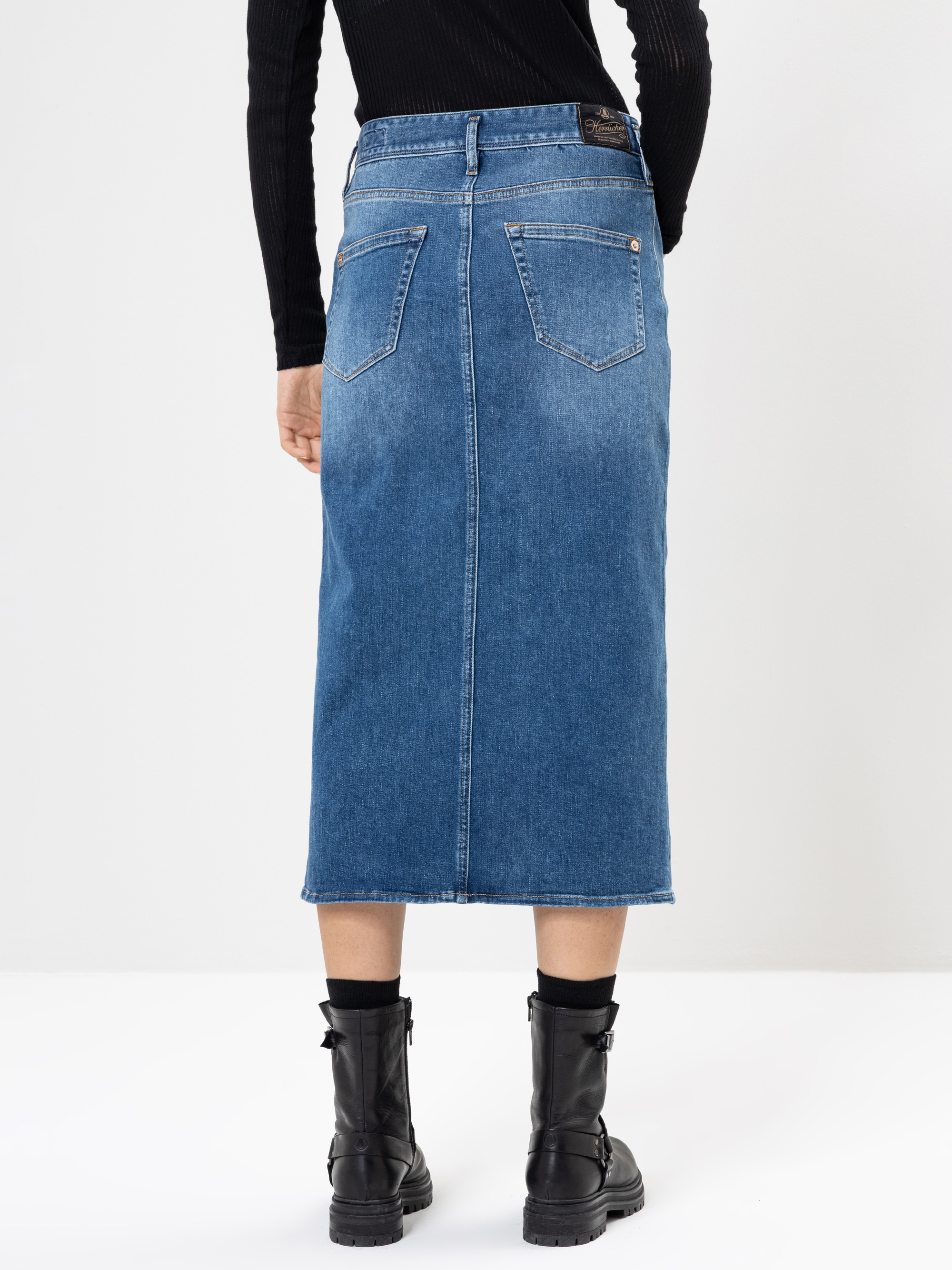 Eine Person wird von hinten gezeigt, die ein schwarzes langärmeliges Rippenoberteil, einen hochtaillierten blauen Jeans-Midirock mit Gesäßtaschen und schwarze Schnürstiefel trägt. Der Hintergrund ist einfarbig und hell.