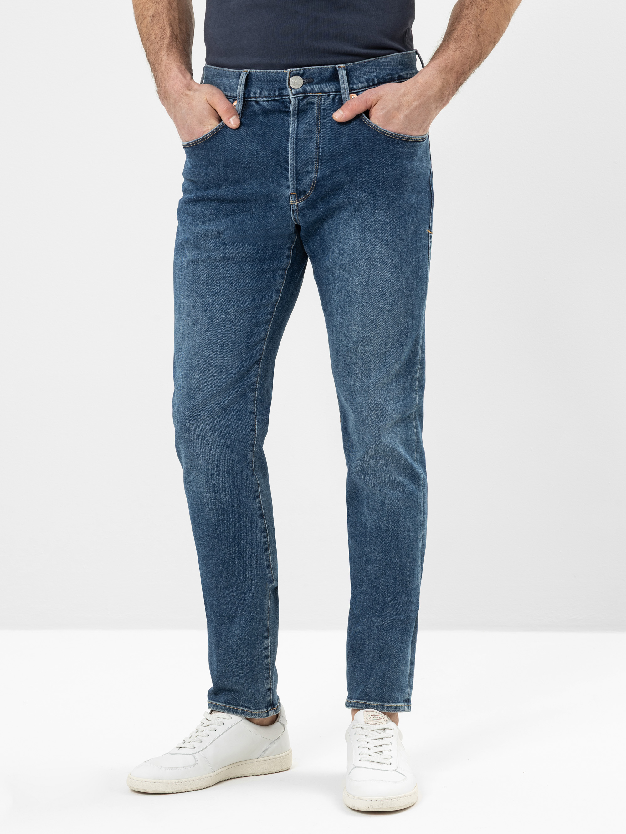 Eine Person, die ein dunkles Hemd, blaue Slim-Fit-Jeans und weiße Turnschuhe trägt, steht mit den Händen in den Taschen vor einem schlichten weißen Hintergrund.
