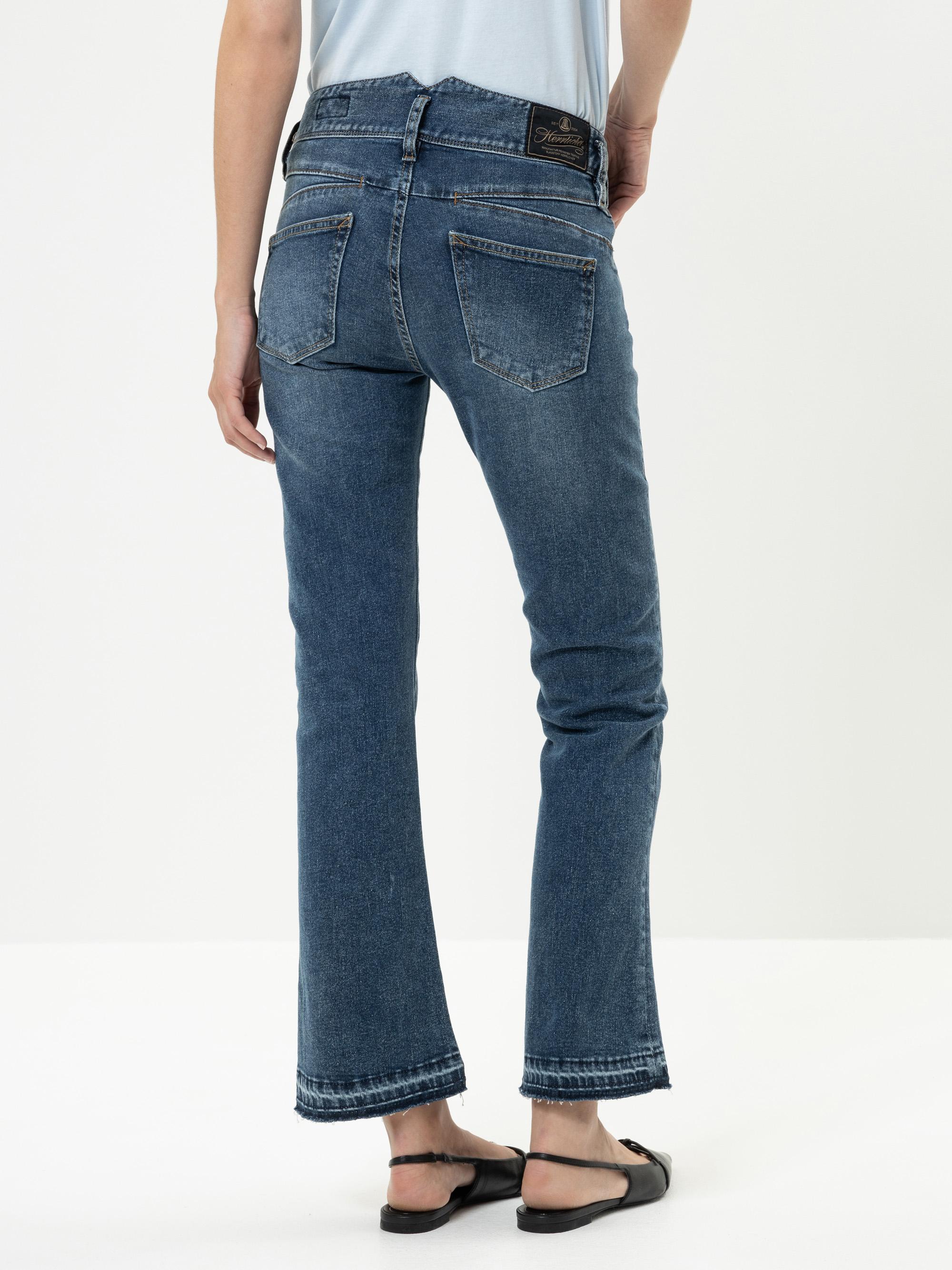 Eine Person in mittelblauen Bootcut-Jeans und schwarzen Sandalen steht mit dem Rücken zur Kamera vor einem schlichten weißen Hintergrund.