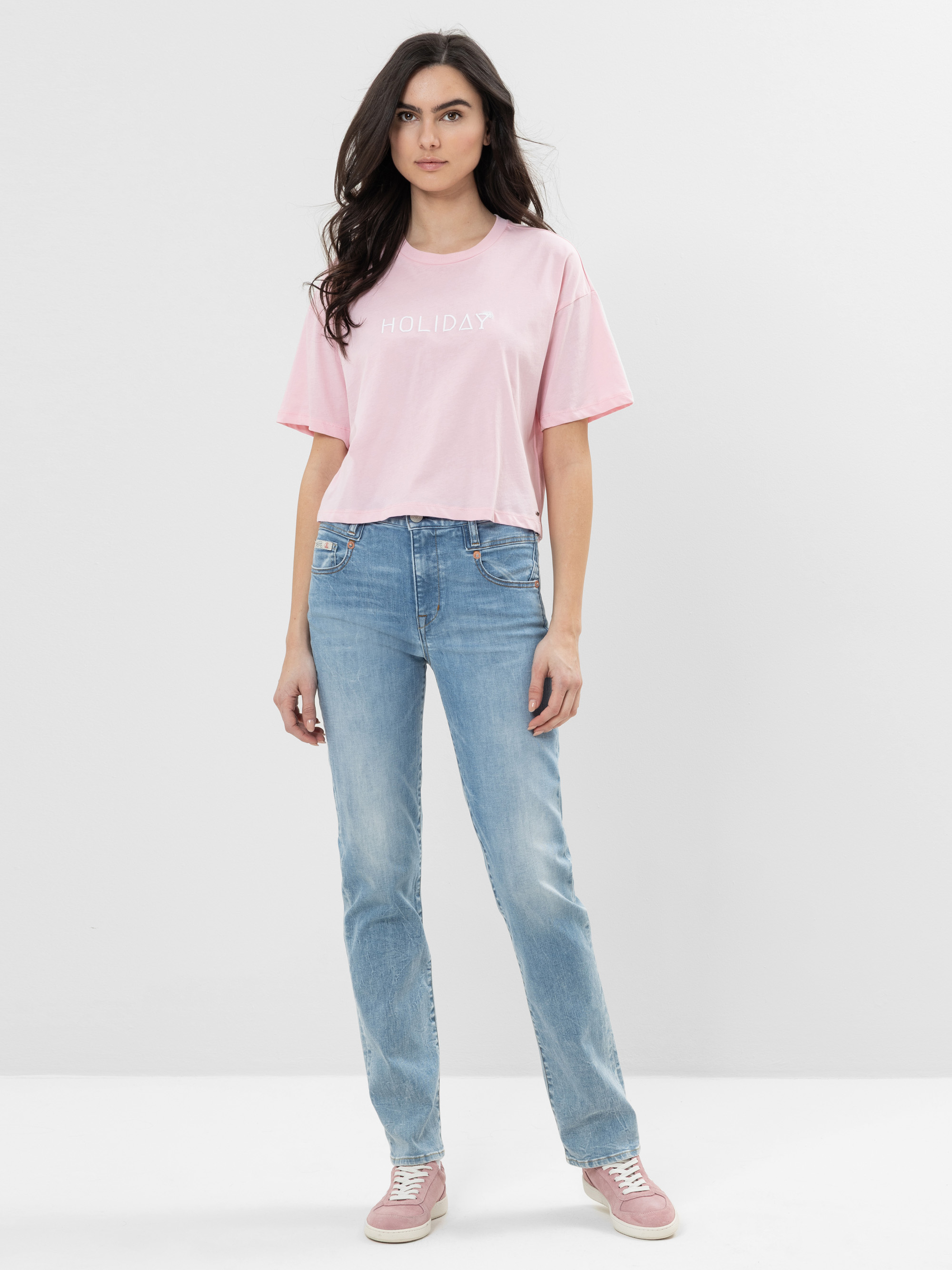 Eine Frau mit langen dunklen Haaren trägt ein hellrosa HOLIDAY-T-Shirt, blaue Jeans und rosa Turnschuhe und steht vor einem schlichten weißen Hintergrund.