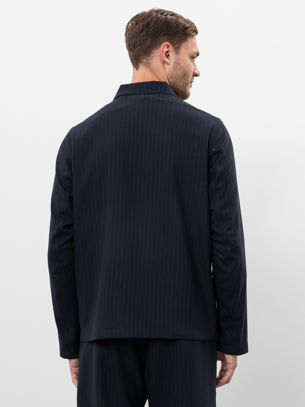 Ein Mann mit kurzen braunen Haaren, von hinten gesehen, trägt eine dunkle marineblaue Nadelstreifen-Anzugsjacke und eine passende Hose vor einem schlichten weißen Hintergrund.