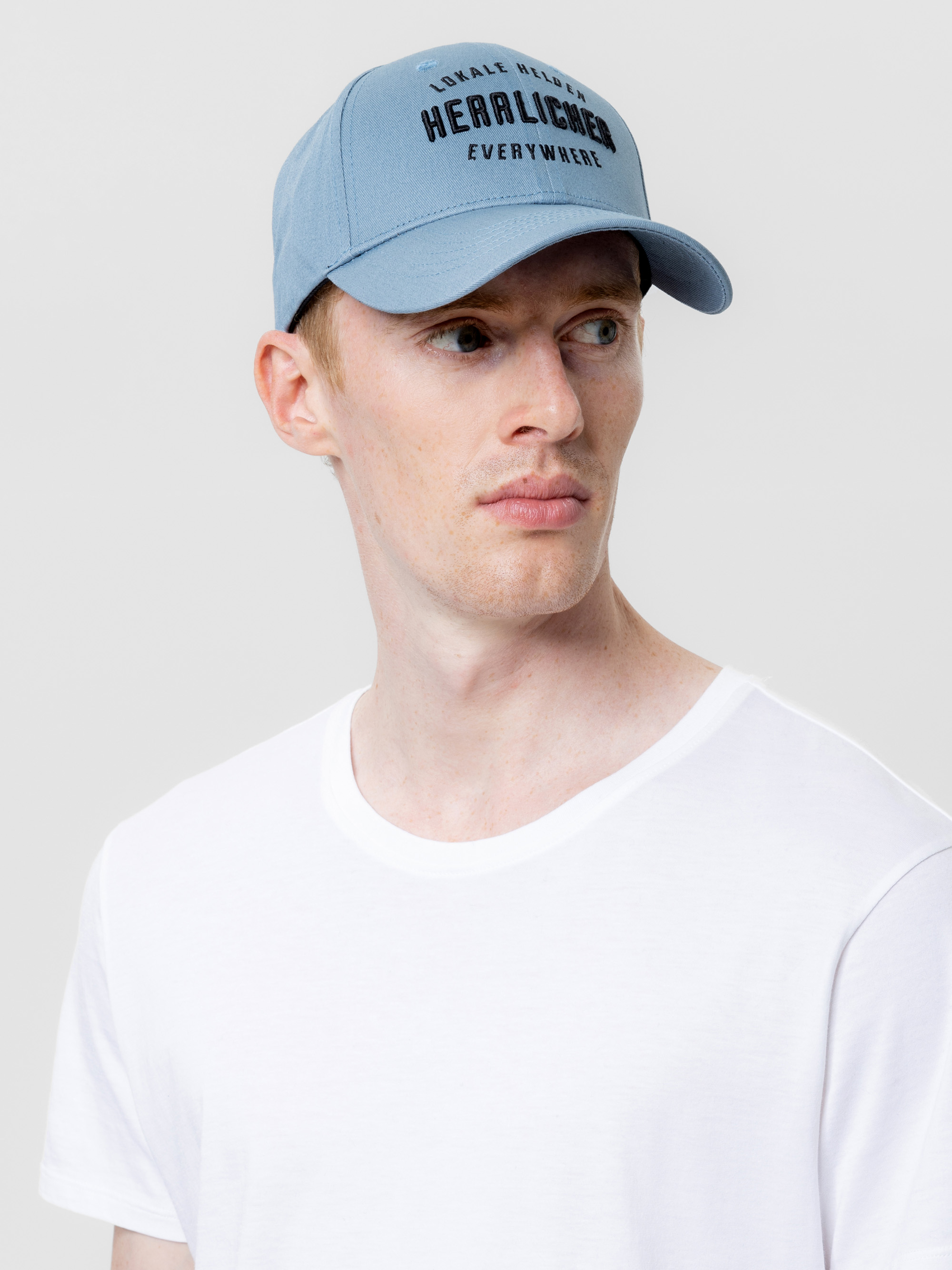 Ein Mann, der eine hellblaue Baseballkappe mit schwarzer Aufschrift und ein schlichtes weißes T-Shirt trägt, schaut vor einem schlichten hellgrauen Hintergrund leicht nach links.