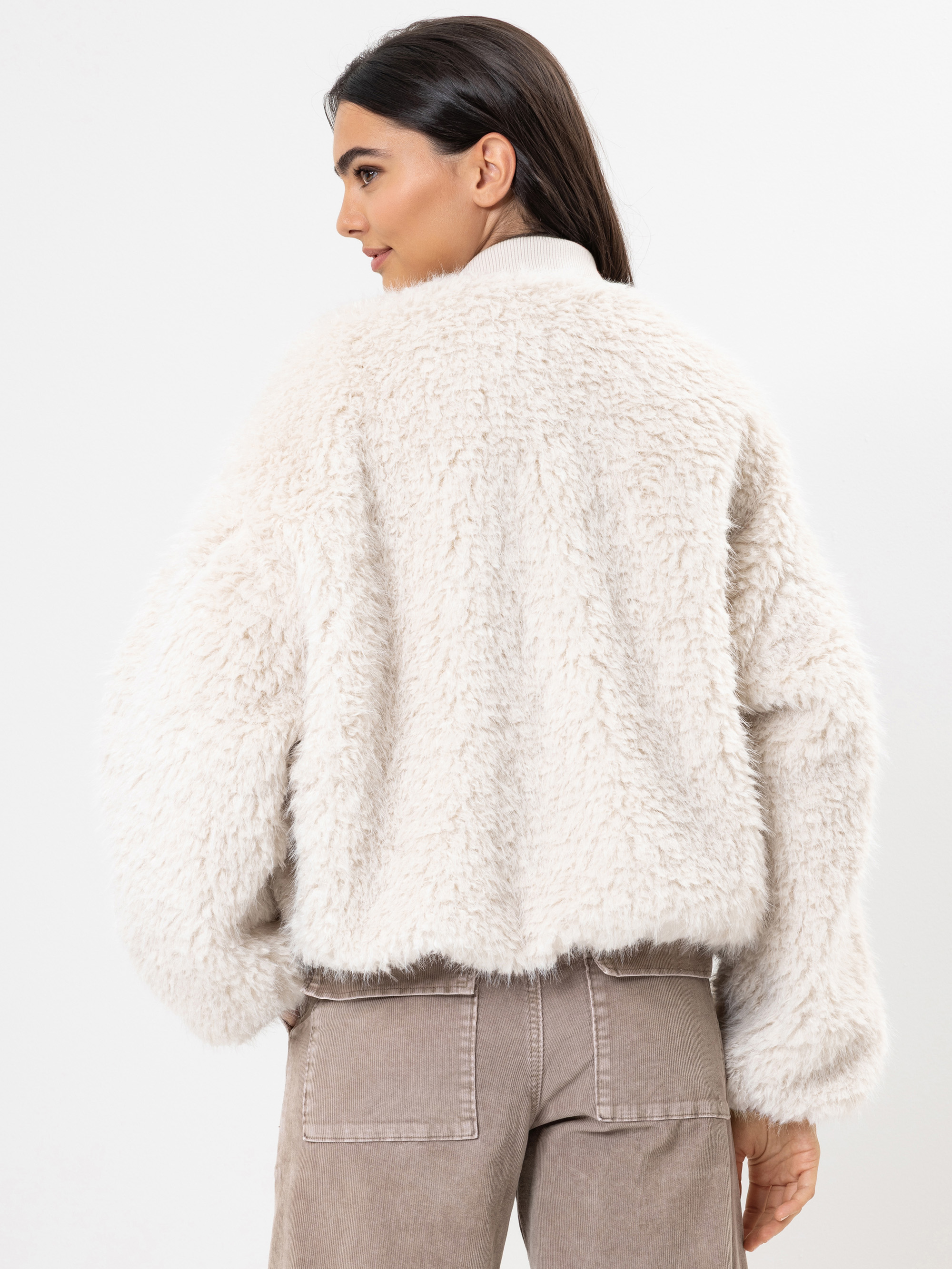 Eine Frau mit langen dunklen Haaren steht mit dem Gesicht zur Wand und trägt eine flauschige, wollweiße Kunstpelzjacke und eine taupefarbene Hose vor einem schlichten weißen Hintergrund.
