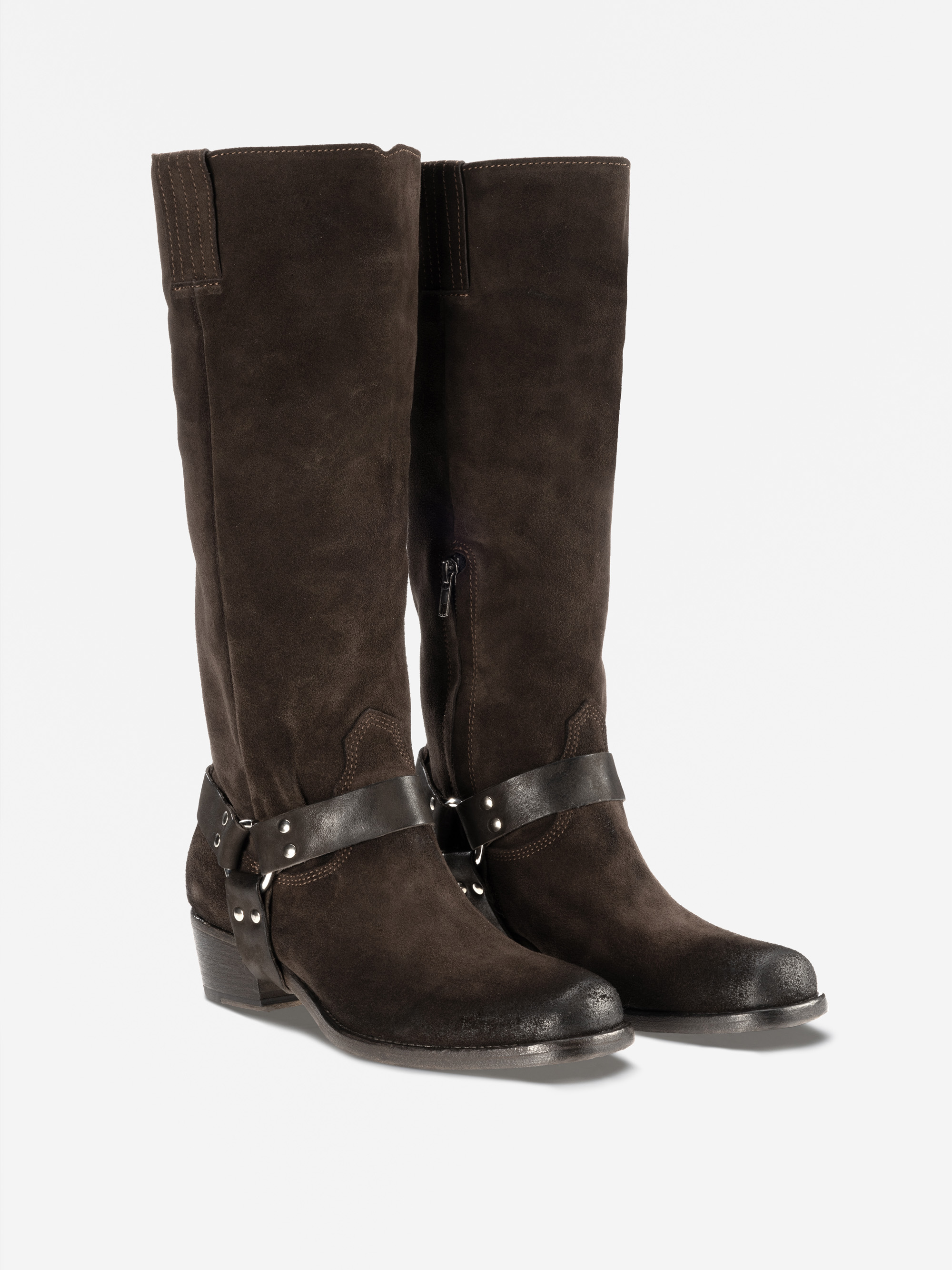 Ein Paar dunkelbraune, halbhohe Wildlederstiefel mit schwarzem Stöckelabsatz, schwarzen Lederriemen mit Metallnieten um die Knöchel und seitlichen Reißverschlüssen auf weißem Hintergrund.