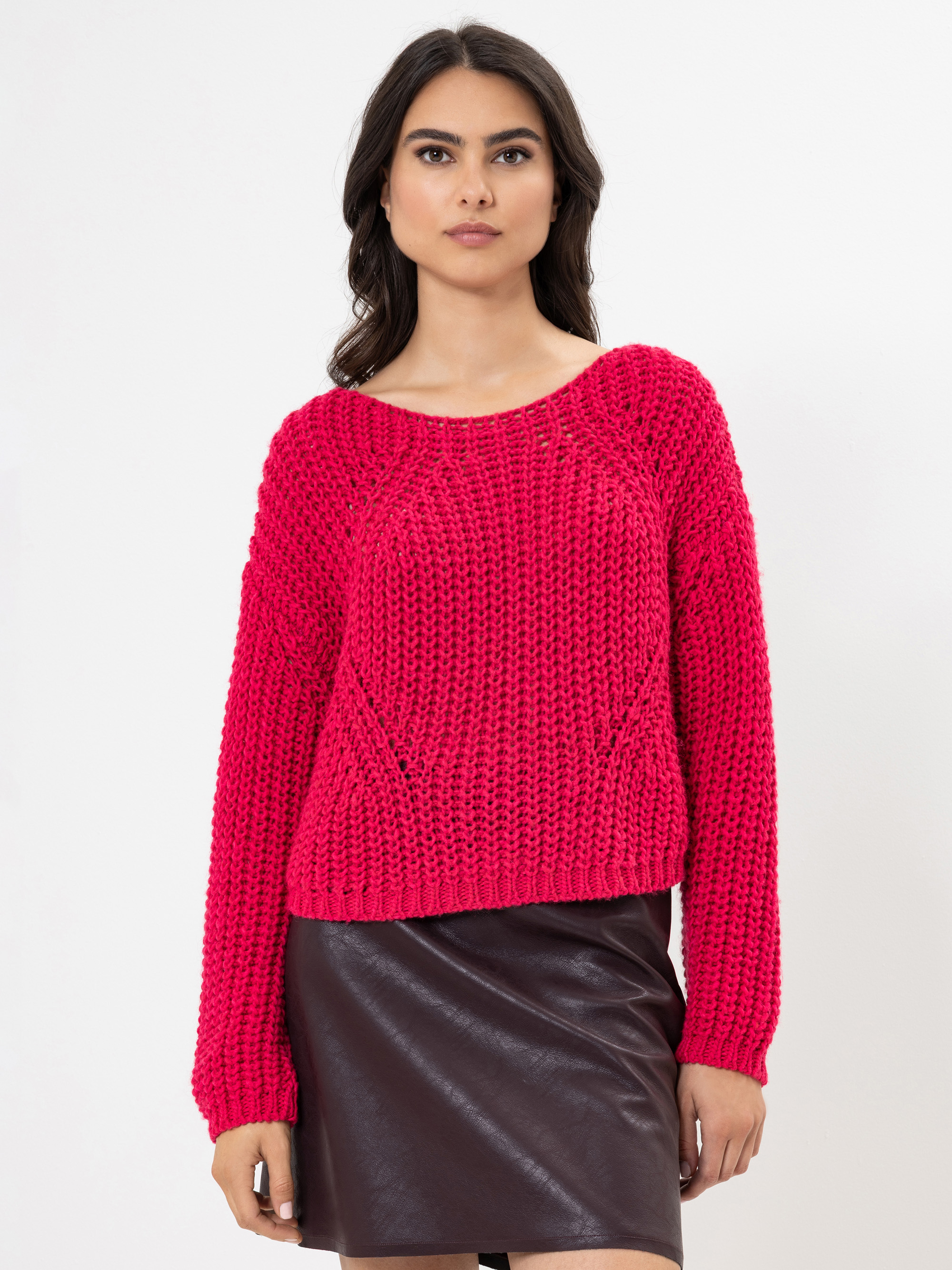 Eine Frau, die einen hellrosa Strickpullover und einen dunkelbraunen Lederrock trägt, steht vor einem schlichten weißen Hintergrund und blickt mit neutraler Miene nach vorn.
