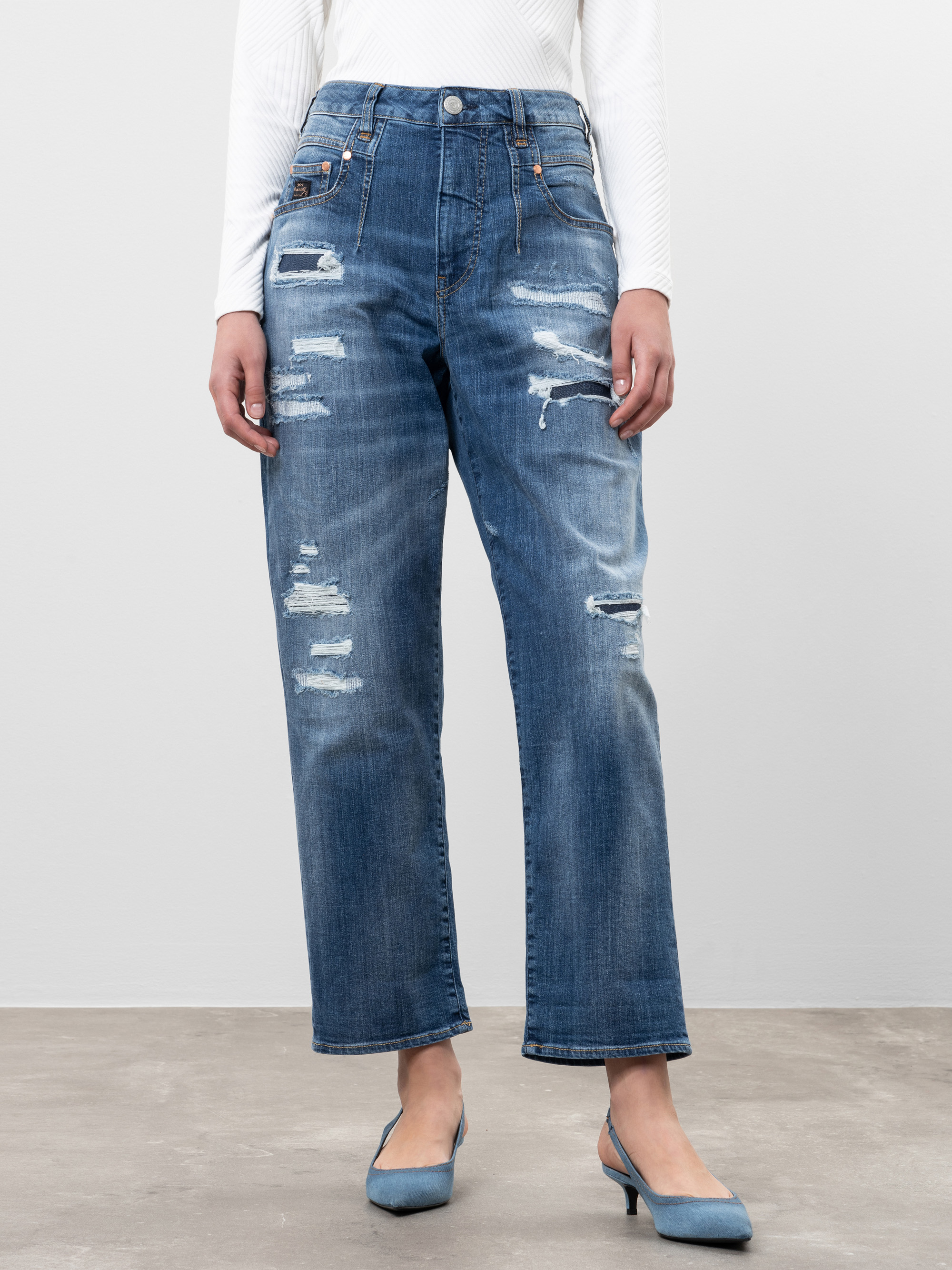 Eine Person, die ein weißes langärmeliges Oberteil, eine blaue Jeans mit geraden Beinen und Rissen und hellblaue Schuhe mit Absätzen trägt, steht auf einem hellen Boden vor einem einfarbigen Hintergrund.