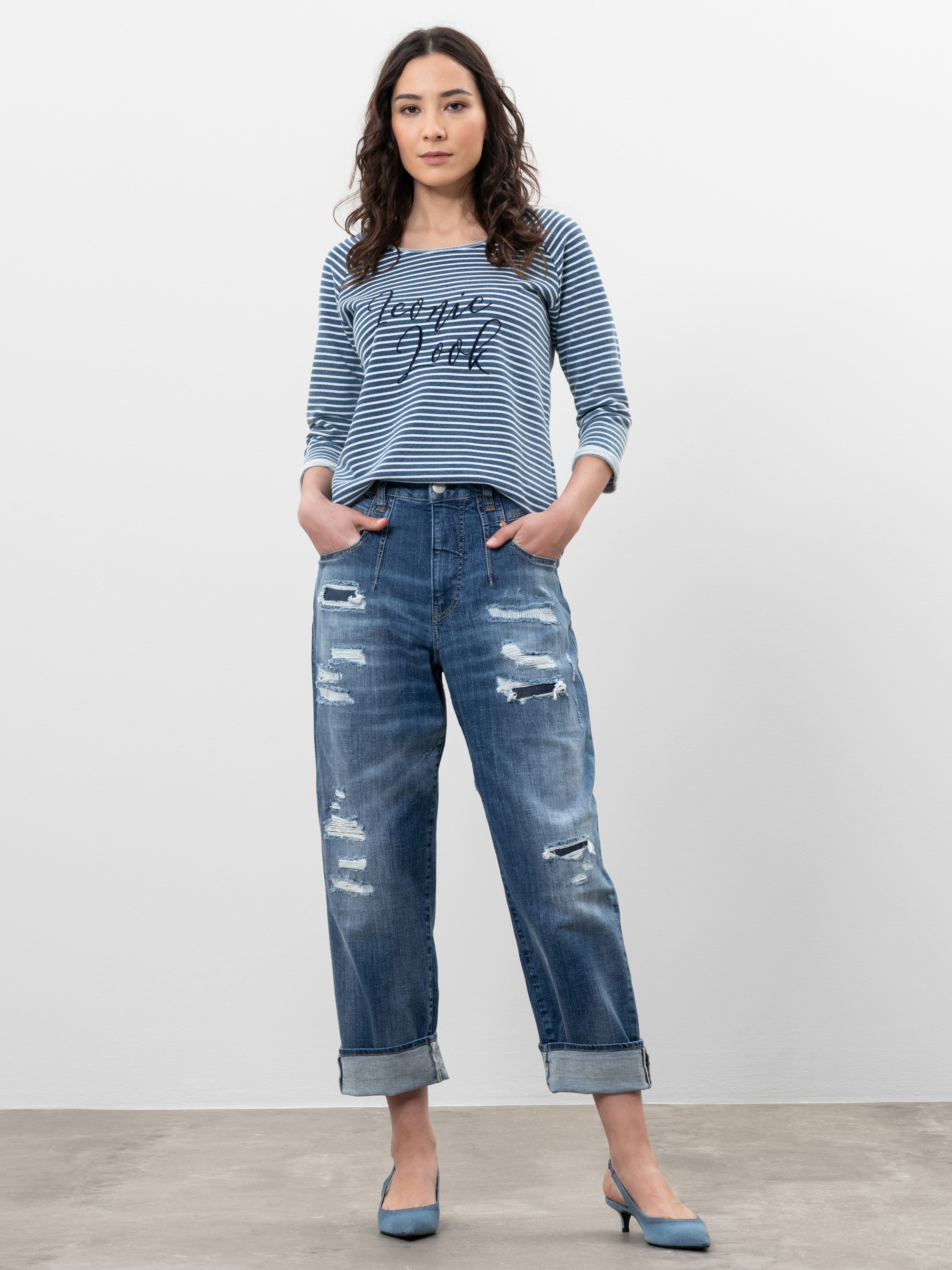Eine Frau steht vor einem schlichten Hintergrund und trägt ein blau-weiß gestreiftes, langärmeliges Oberteil mit der Aufschrift "Bonjour Paris", zerschlissene, aufgerollte Jeans und blaue Absätze. Ihre Hände stecken in den Taschen und sie hat einen n